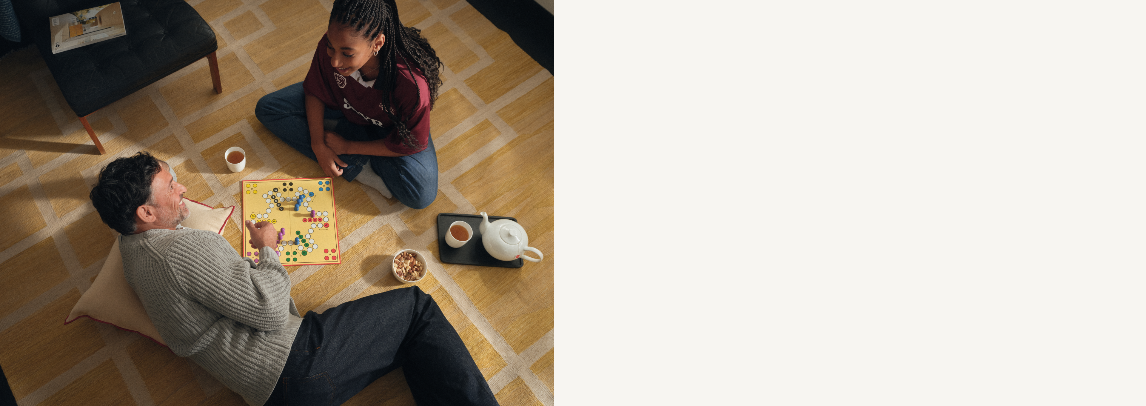 Ein lächelnder Mann mit Brille sitzt auf einem Kissen und spielt ein Brettspiel mit bunten Spielfiguren. In der Nähe steht eine Tasse Tee auf einem gemusterten Teppich.