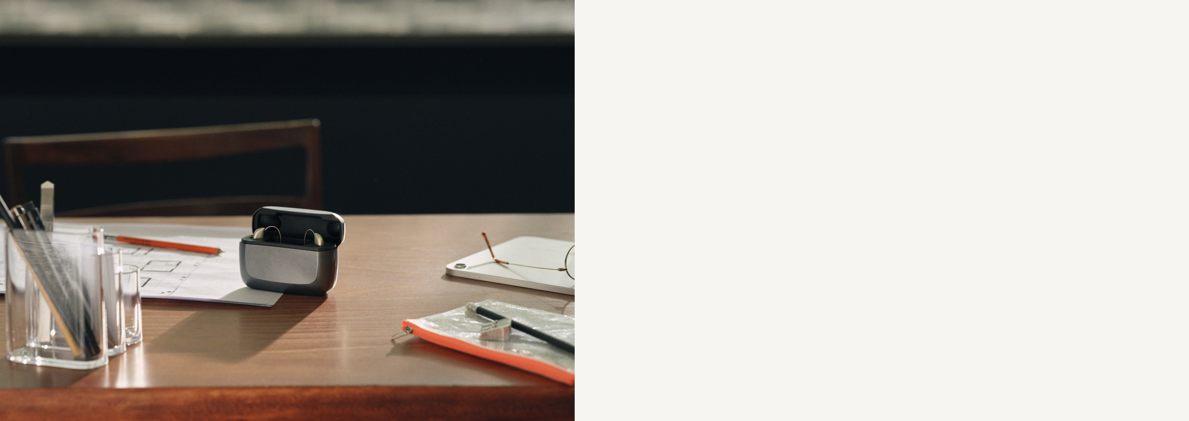 Ein Paar kabellose Ohrhörer in einem offenen Ladeetui auf einem Holztisch, umgeben von Papieren, einem Bleistift und einem transparenten Stiftehalter.