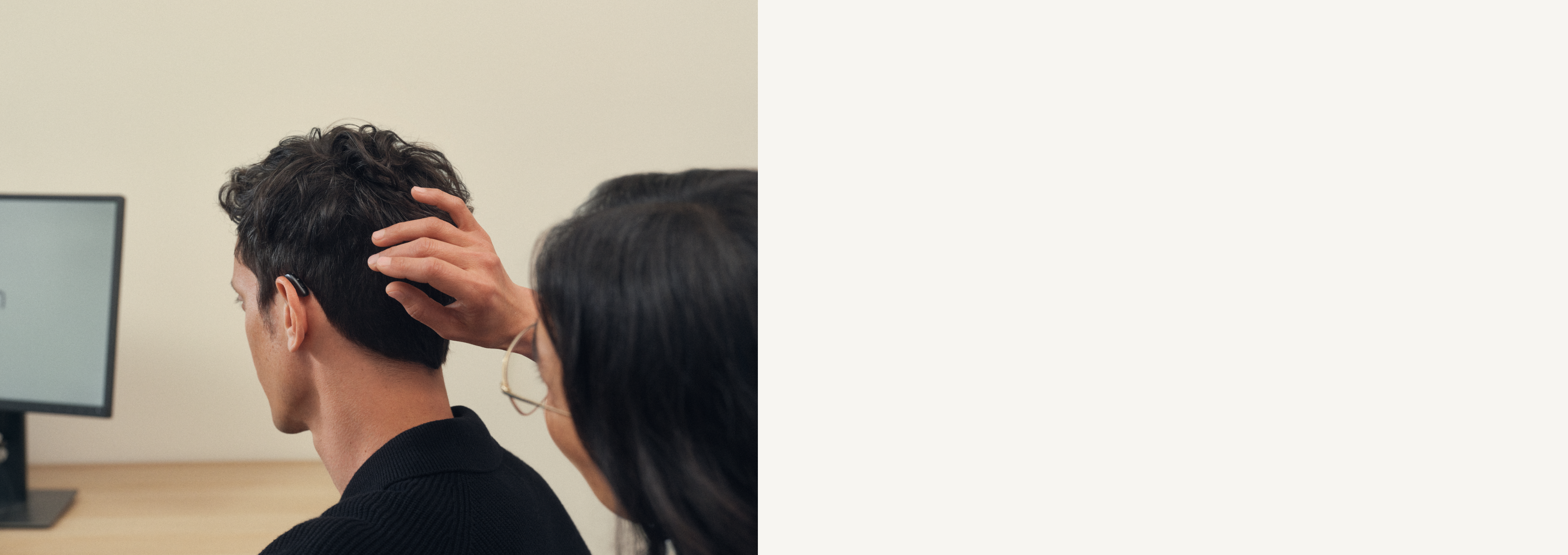 Eine Person passt ein Hörgerät am Ohr eines Mannes an, während dieser in einem Büro vor einem Computerbildschirm sitzt.