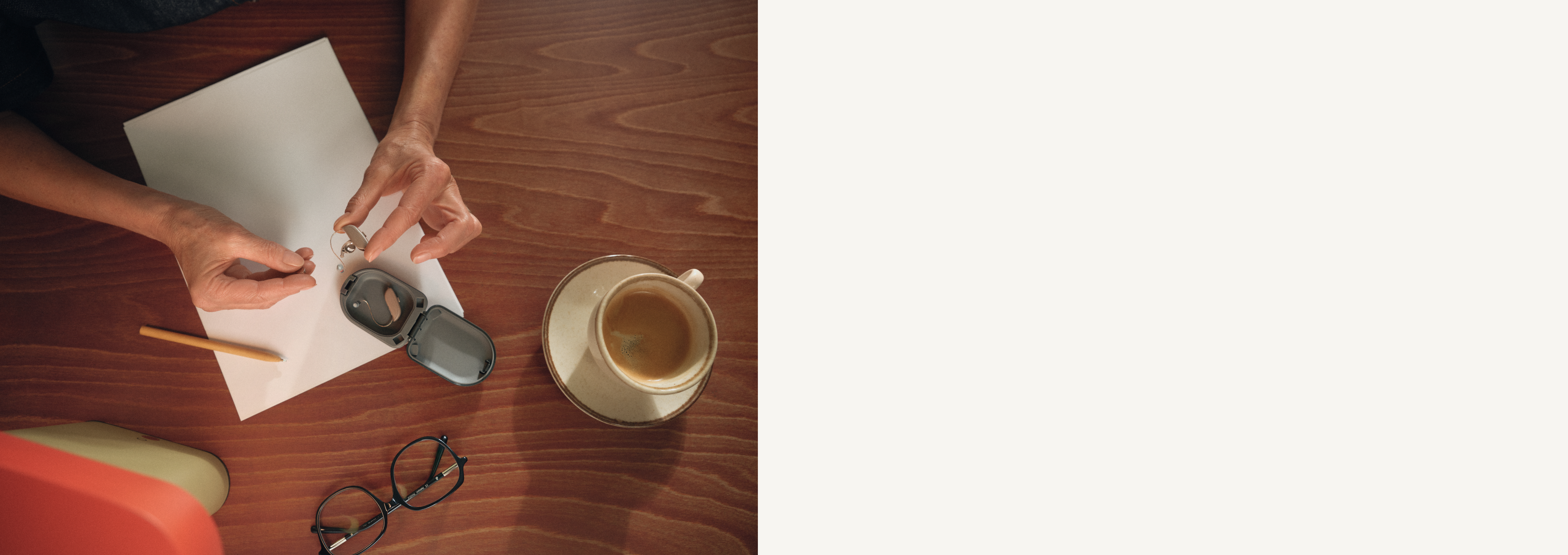 Hände, die einen kleinen Gegenstand auf einem Holztisch halten, auf dem sich ein leeres Blatt Papier, ein Bleistift, eine Brille, ein offenes Etui und eine Tasse Kaffee befinden.