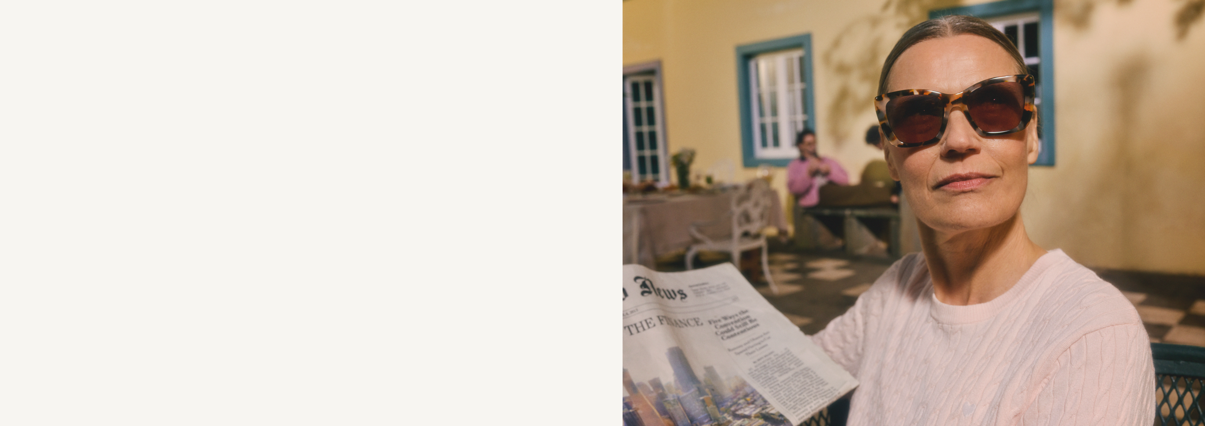 Femme portant des lunettes de soleil et tenant un journal, assise à la terrasse d'un café, avec un homme en arrière-plan.