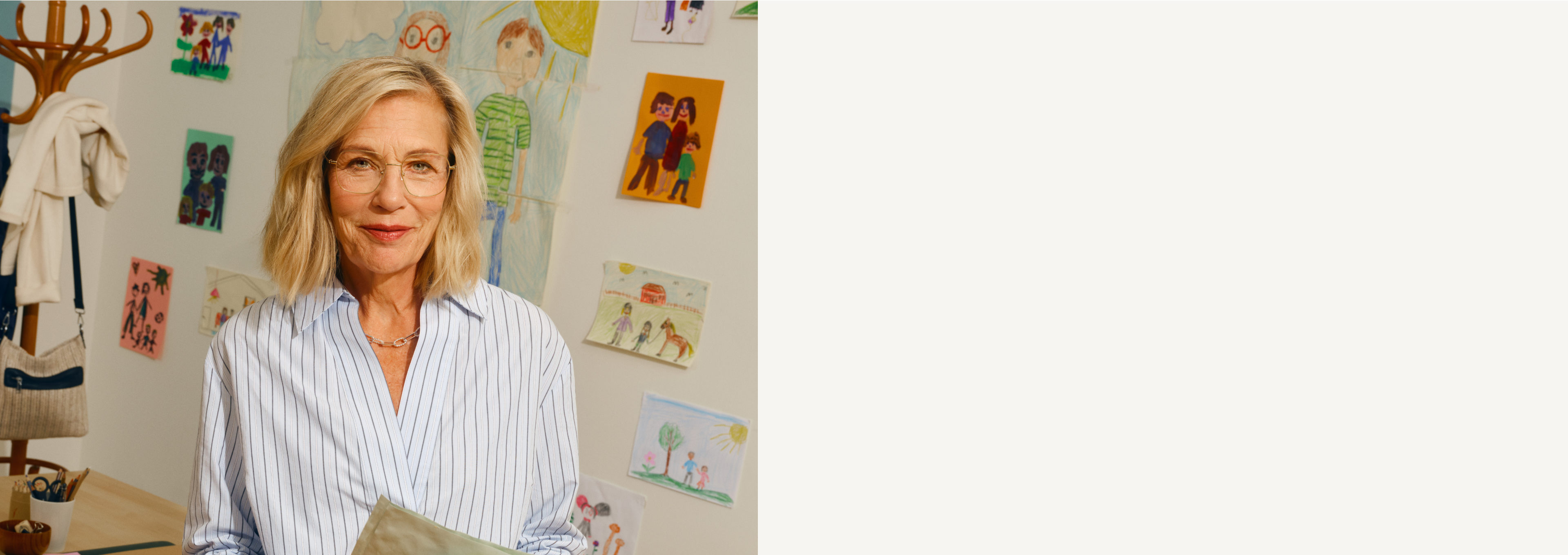 Lächelnde Frau mit Brille steht in einem Raum mit Kinderzeichnungen an der Wand und trägt ein gestreiftes Shirt.