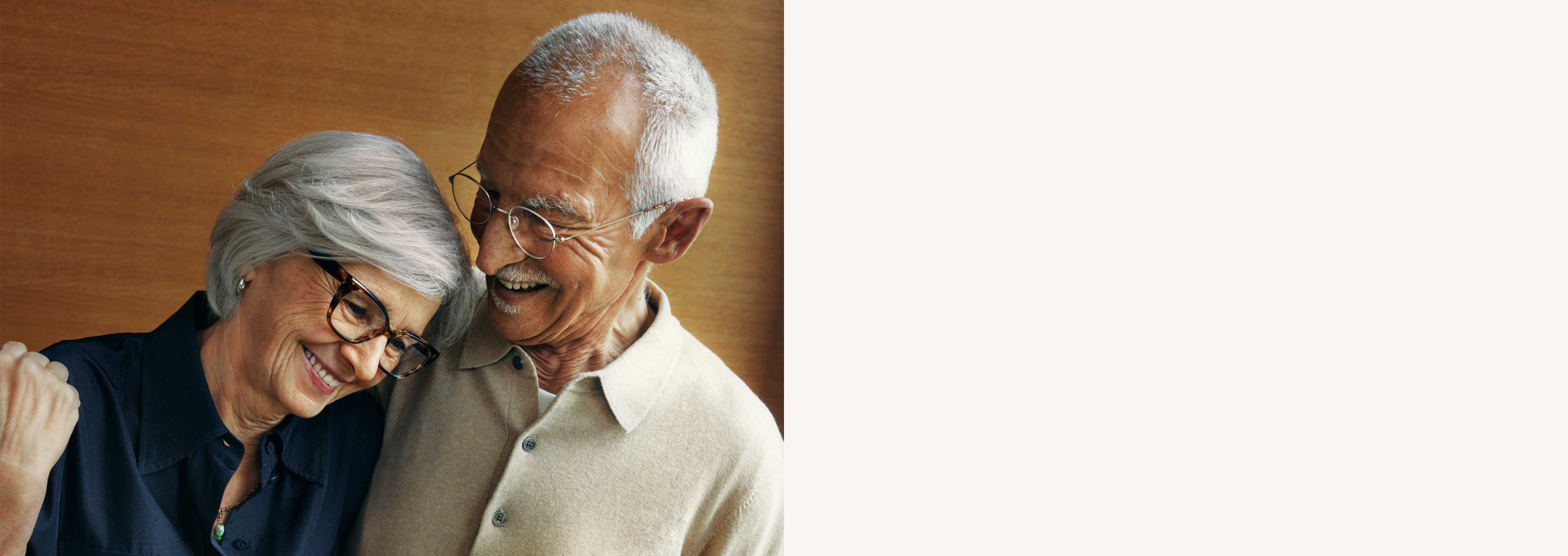 Älteres Paar, das lächelt und Brillen trägt; die Frau lehnt ihren Kopf an die Schulter des Mannes vor einem warmen, hölzernen Hintergrund.