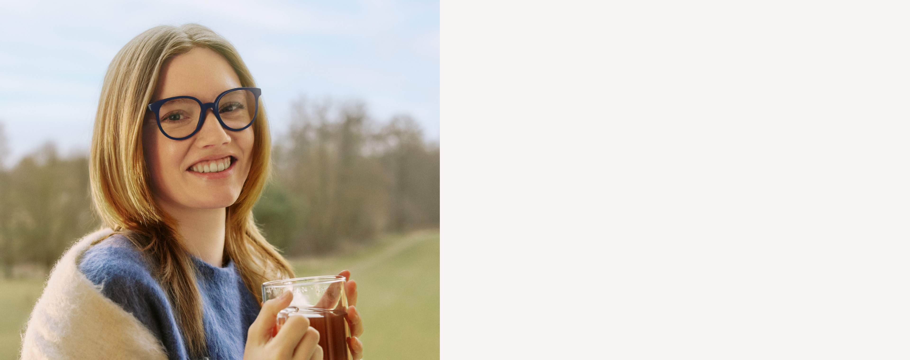 Femme avec des lunettes souriant, tenant une tasse de thé, portant un pull bleu et blanc, avec un arrière-plan flou en extérieur.