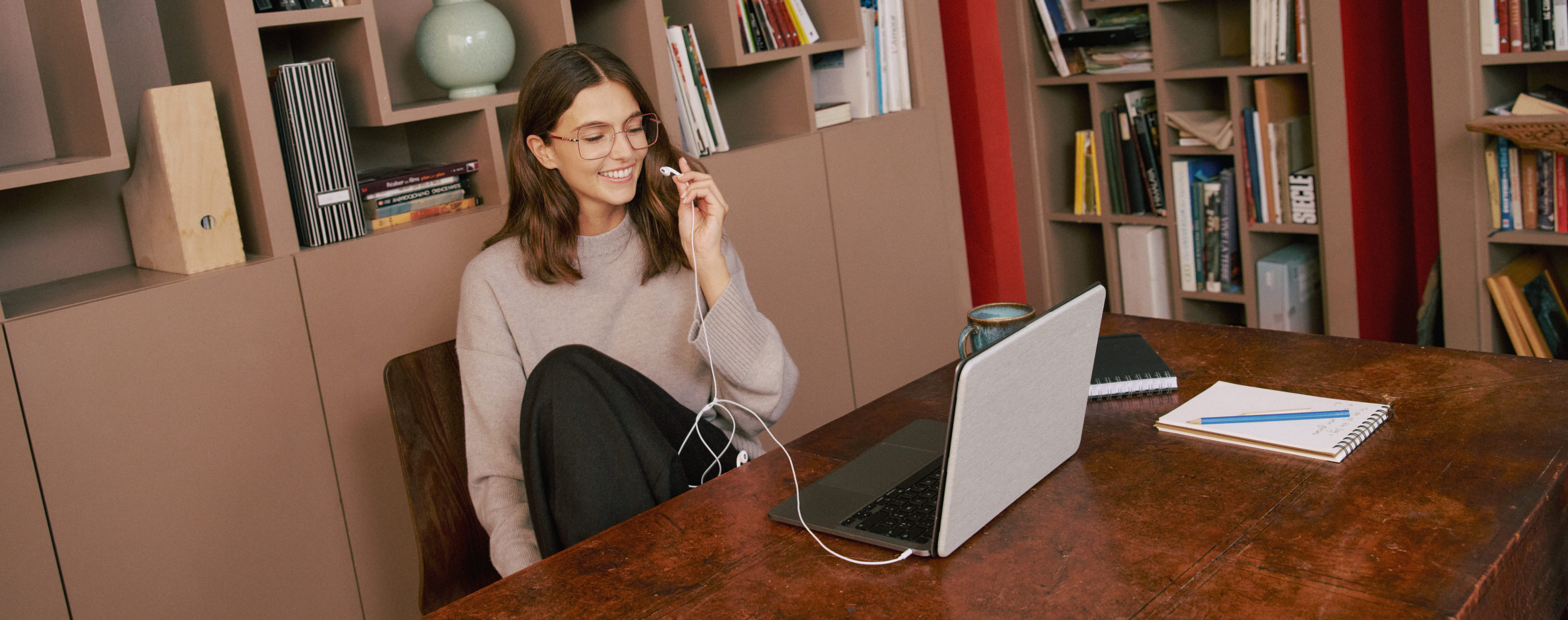 Una donna con gli occhiali è seduta a un tavolo di legno su cui sono appoggiati un computer portatile, delle cuffie, un taccuino, una penna e una tazza, e sorride in una stanza accogliente con delle librerie.