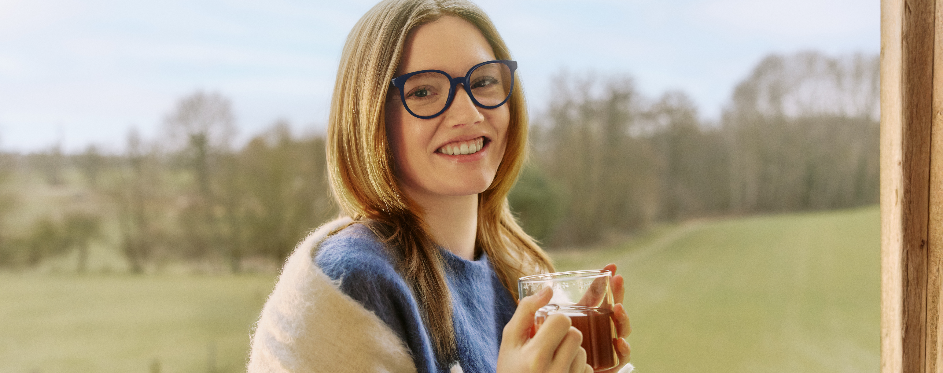 Una donna con gli occhiali sorride mentre tiene in mano una tazza all'aperto.