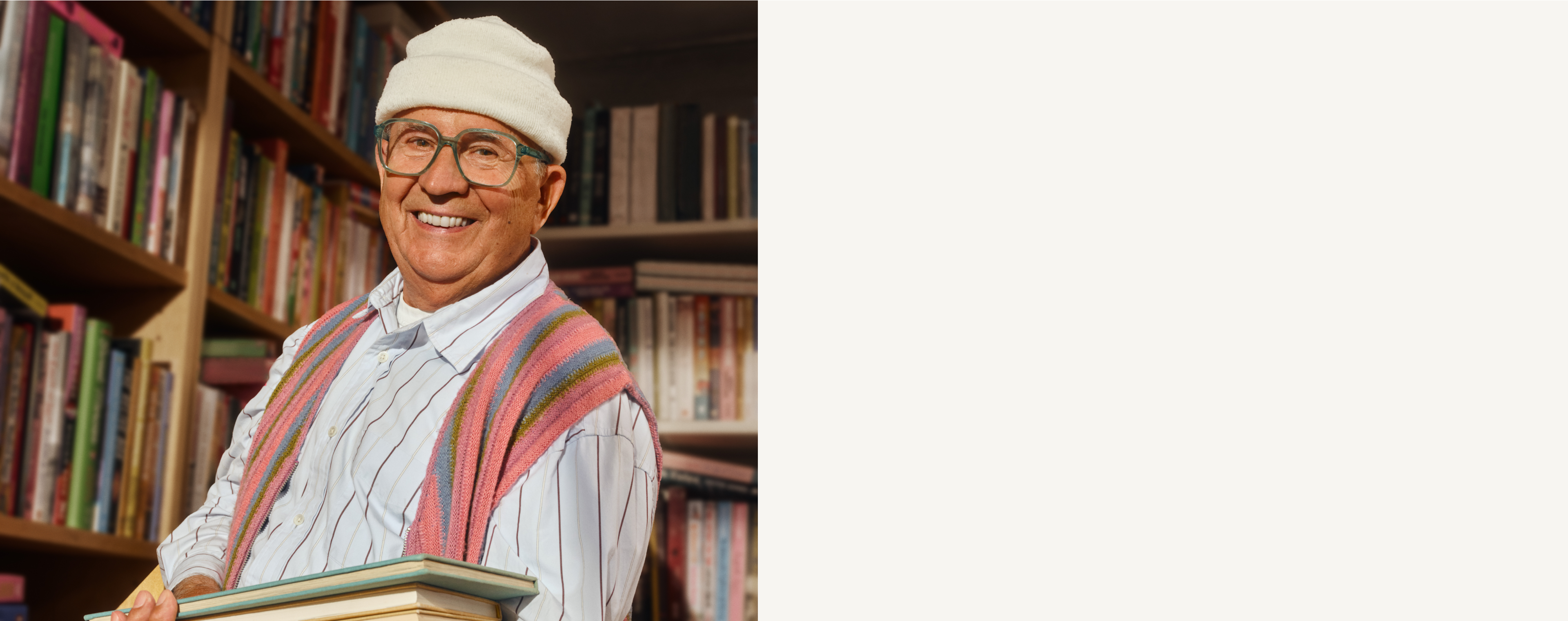 Älterer Herr mit Brille und Mütze, hält Bücher in einer Bibliothek, mit bunten Regalen im Hintergrund.