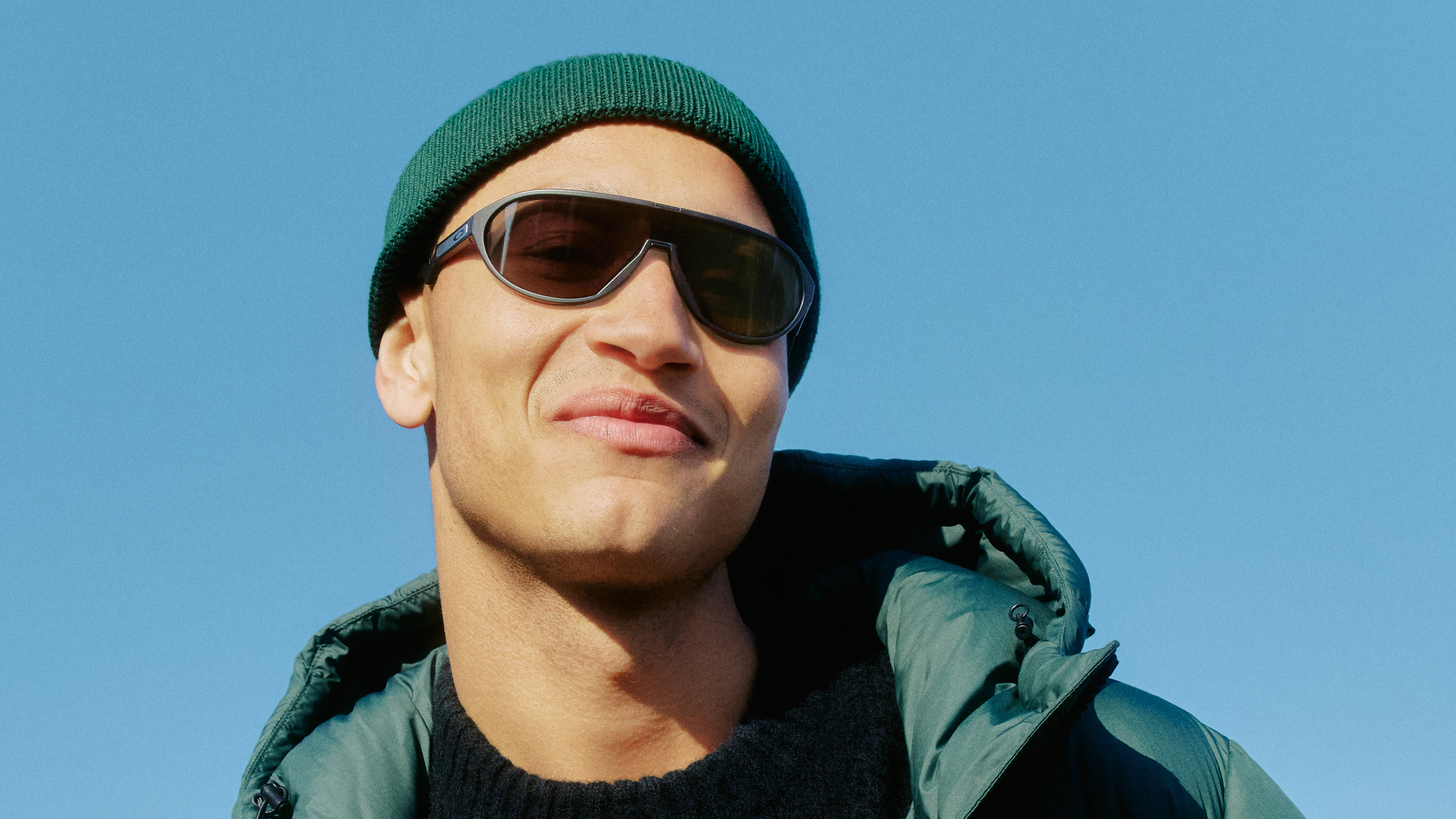 Eine Person mit Sonnenbrille, grüner Mütze und dunkler Jacke lächelt vor einem strahlend blauen Himmel.