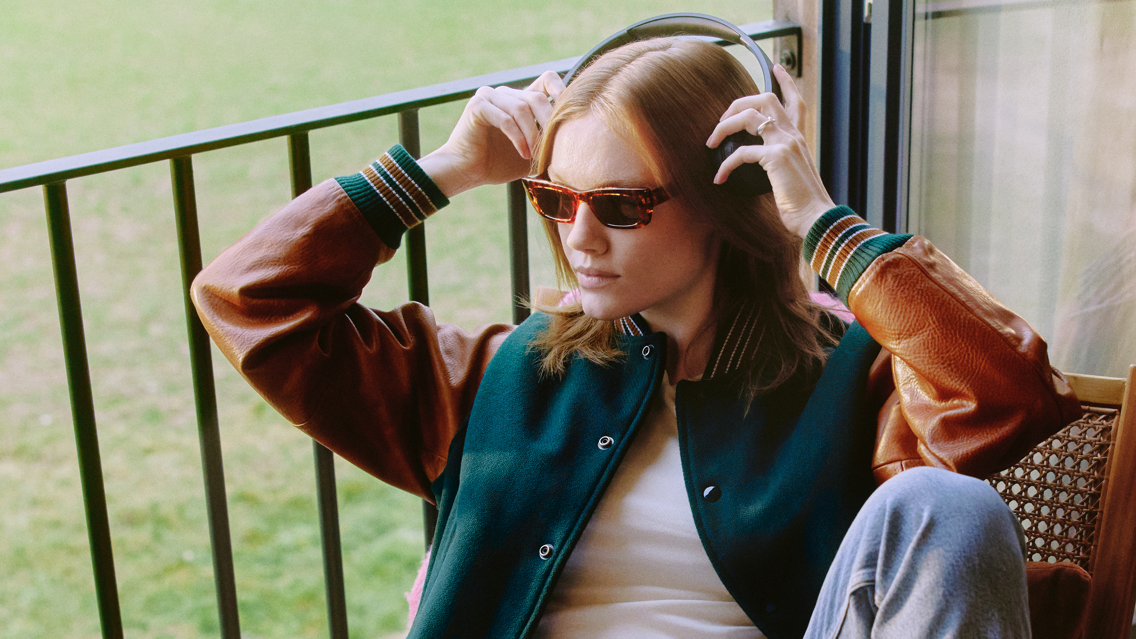 Eine Person mit Sonnenbrille und Kopfhörern, die an einem Fenster sitzt und nach draußen schaut. Sie trägt eine College-Jacke und Jeans.