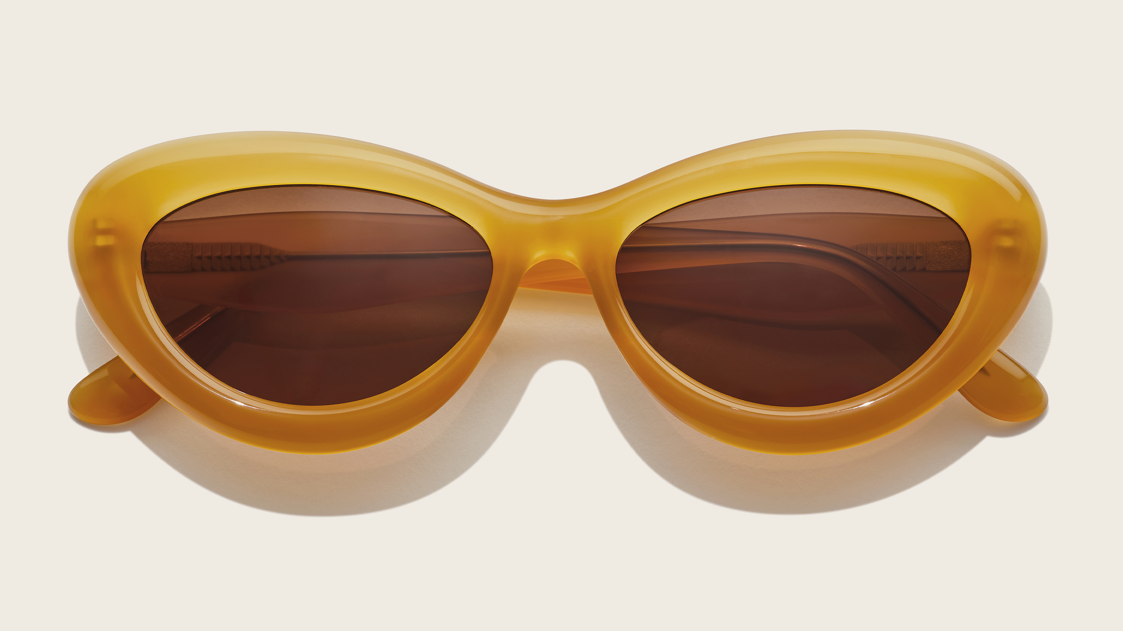 Gelbe Cat-Eye-Sonnenbrille mit braunen Gläsern vor einem hellbeigen Hintergrund.
