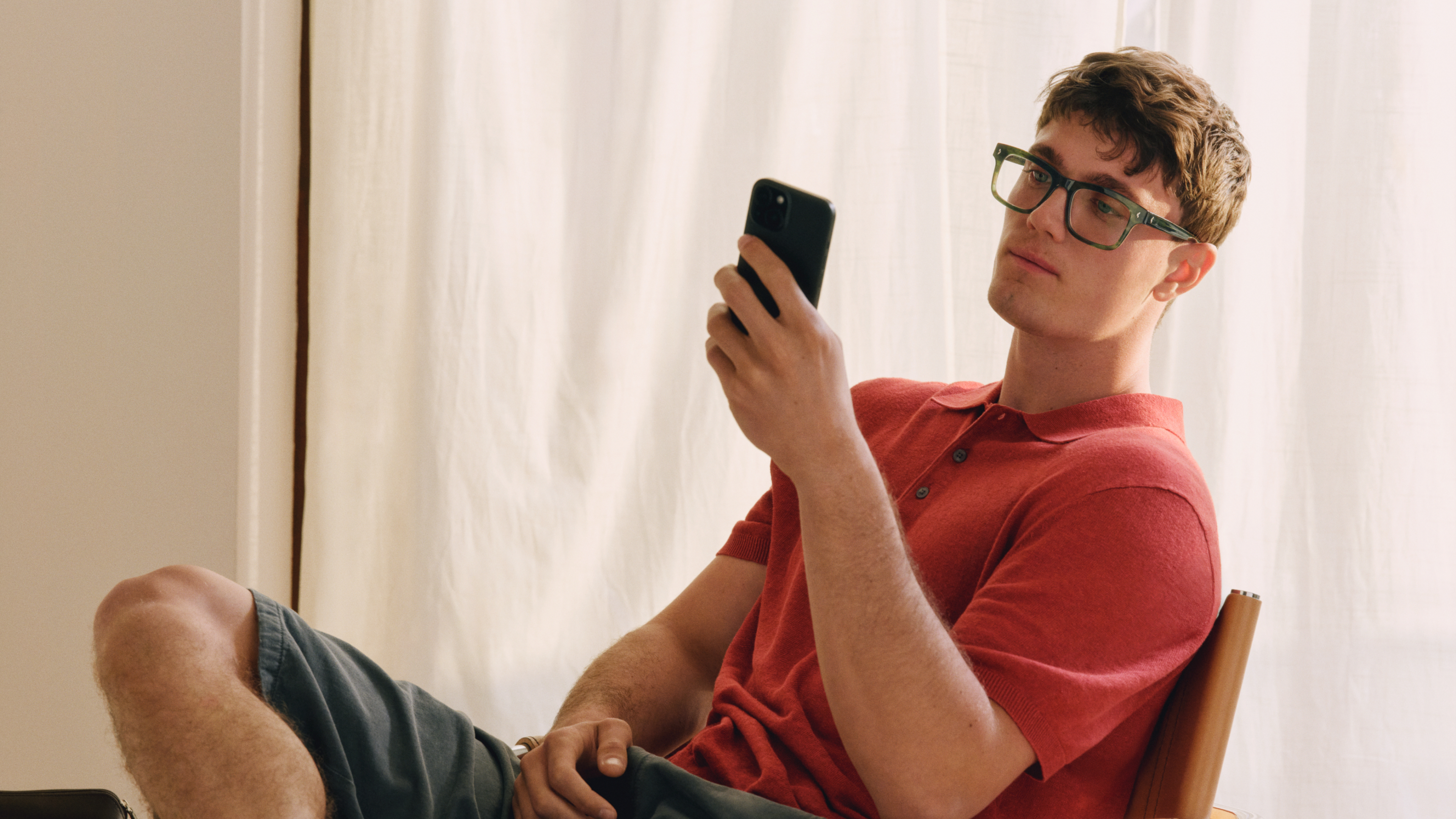 Mann mit Brille, der ein rotes Poloshirt und kurze Hosen trägt, sitzt auf einem Stuhl und blickt mit einem entspannten Gesichtsausdruck auf sein Handy. Hinter ihm befindet sich ein weißer Vorhang.