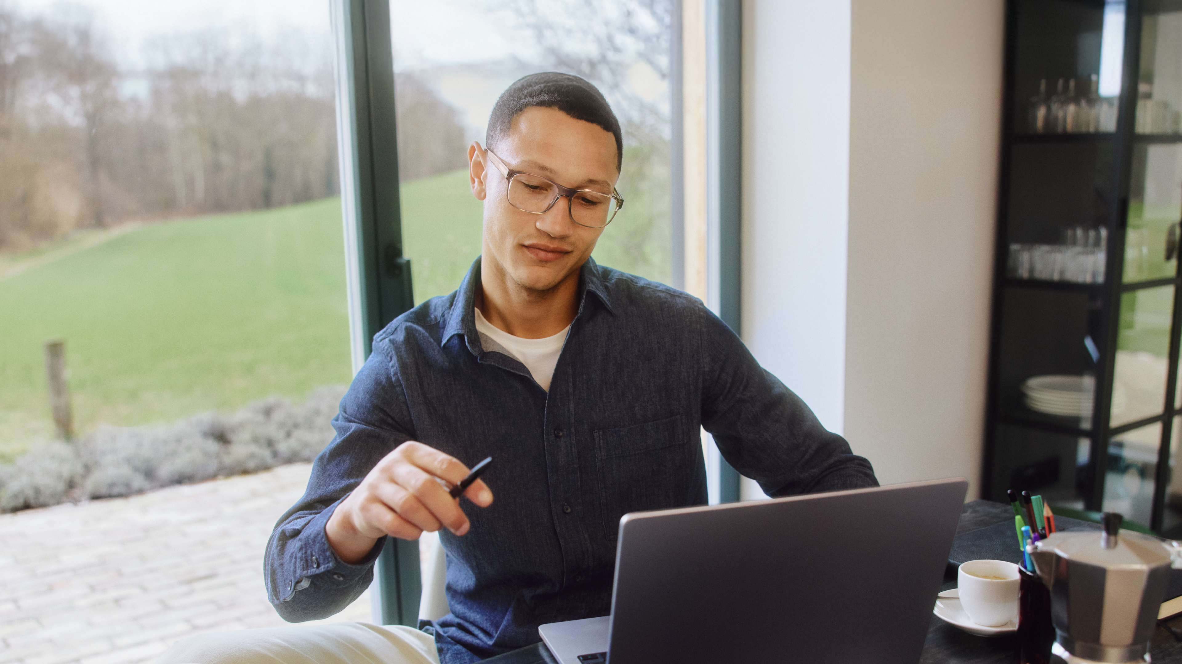 Mann mit Brille, der an einem Tisch in der Nähe großer Fenster an einem Laptop arbeitet und einen Stift in der Hand hält.