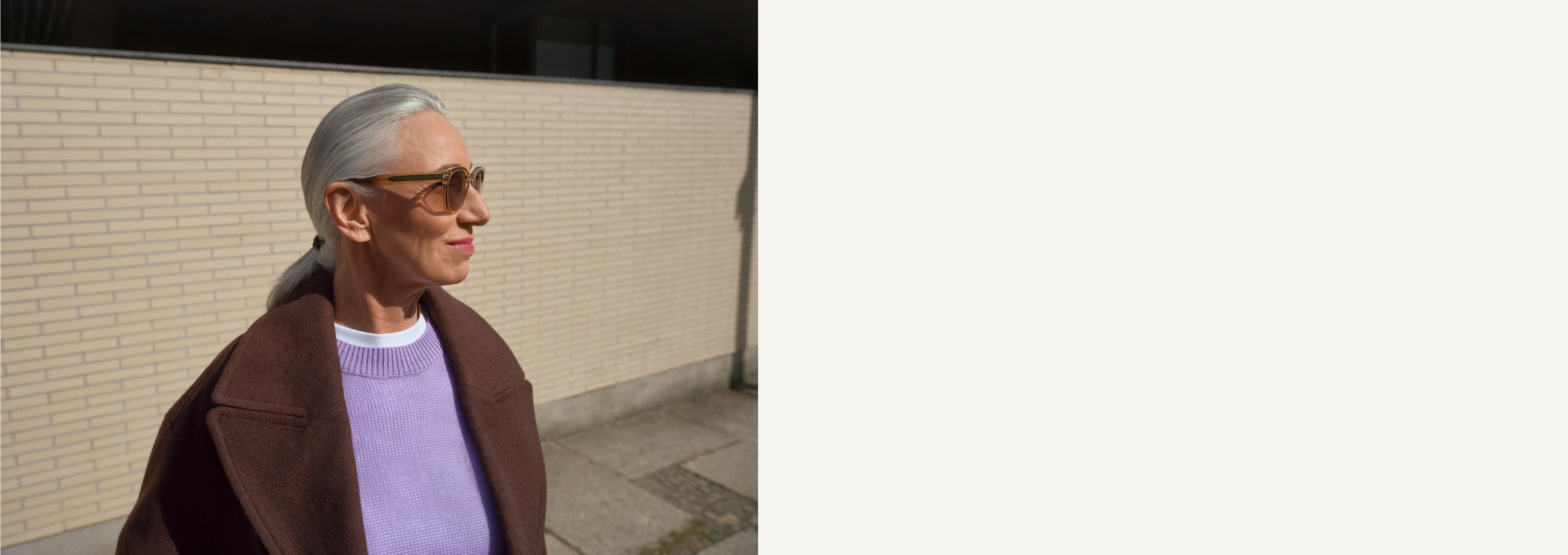 Eine Frau mit grauen Haaren im Pferdeschwanz, Sonnenbrille, braunem Mantel und lila Pullover steht vor einer Ziegelwand und blickt zur Seite.