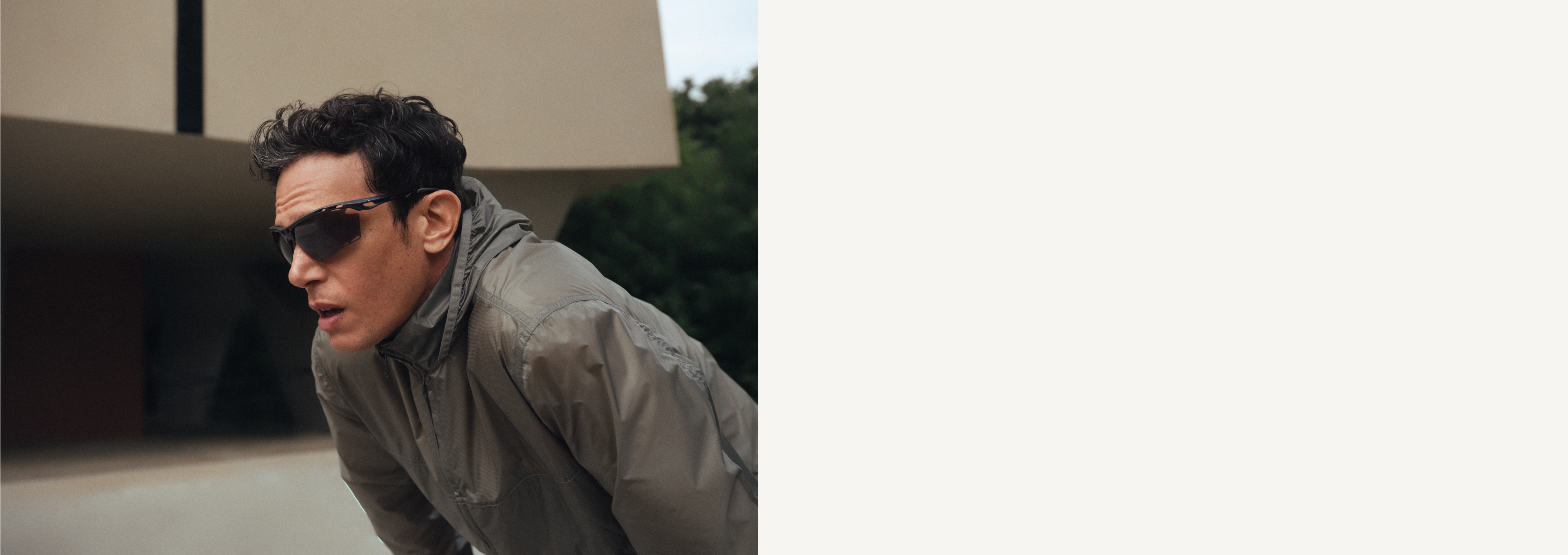 Una persona con giacca grigia e occhiali da sole si china in avanti, con un’espressione concentrata. Sullo sfondo si intravede una struttura architettonica sfocata.