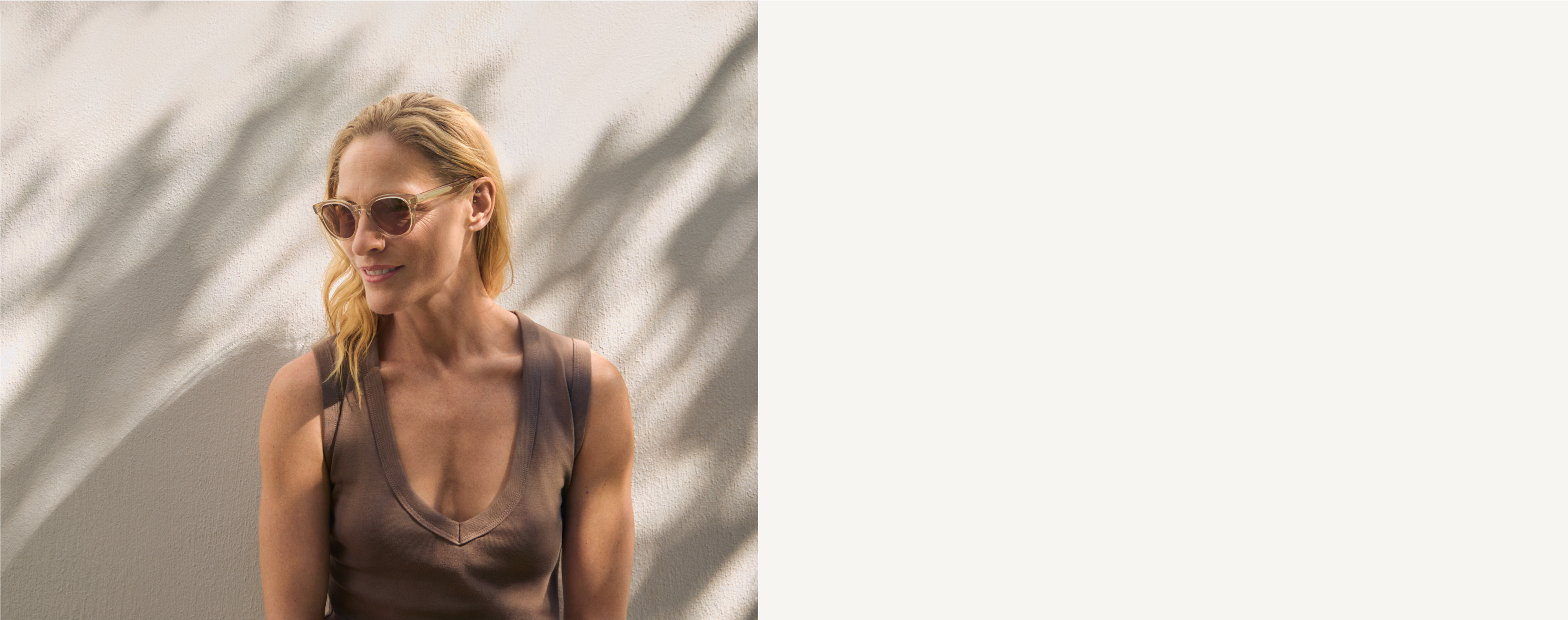Una donna con capelli biondi, occhiali da sole e top senza maniche è in piedi davanti a un muro illuminato dal sole con l'ombra degli alberi.