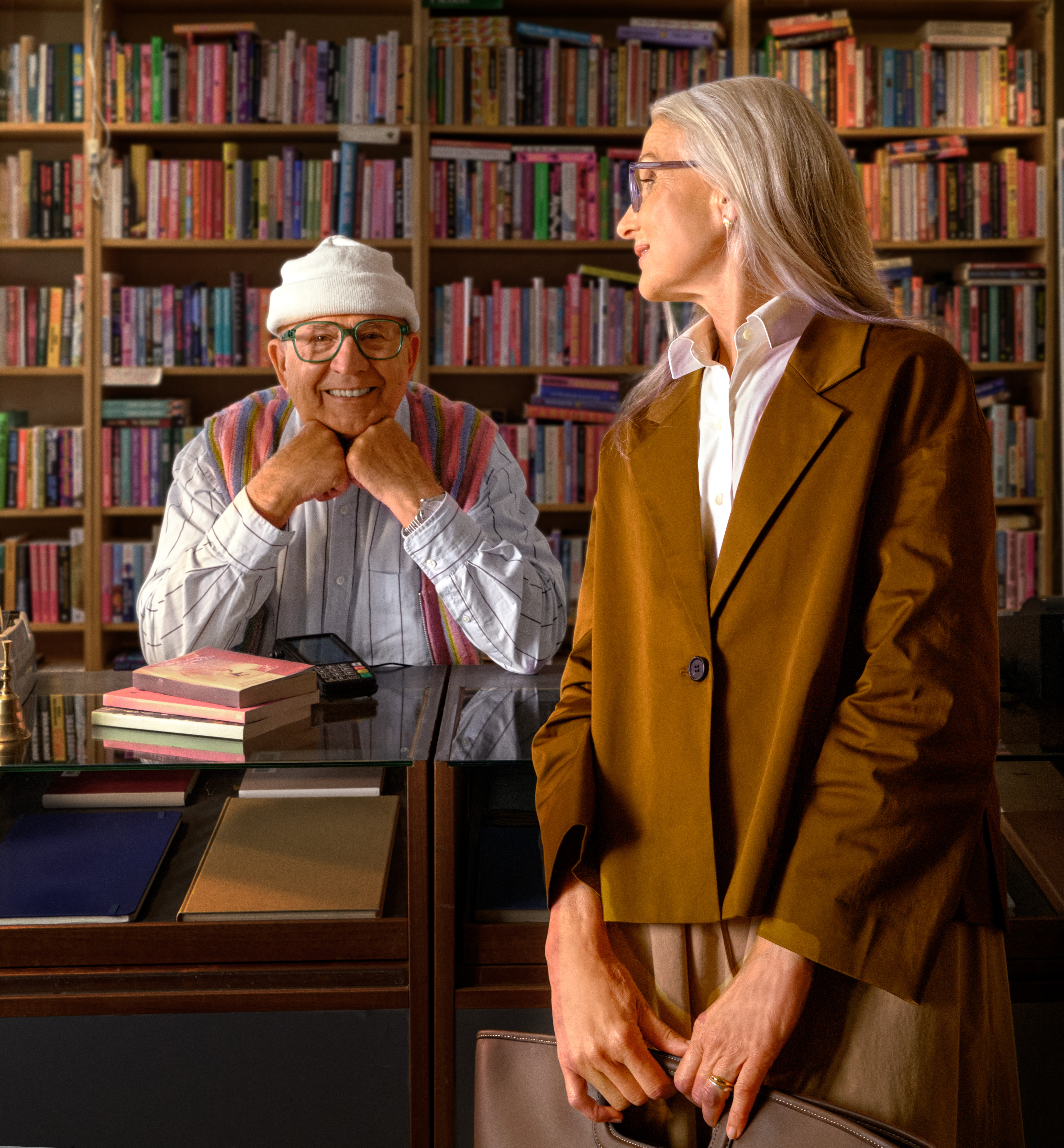Ein älterer Mann und eine Frau stehen in einer gemütlichen Bibliothek. Der Mann lächelt, während die Frau zur Seite blickt. Die Regale sind voller Bücher.
