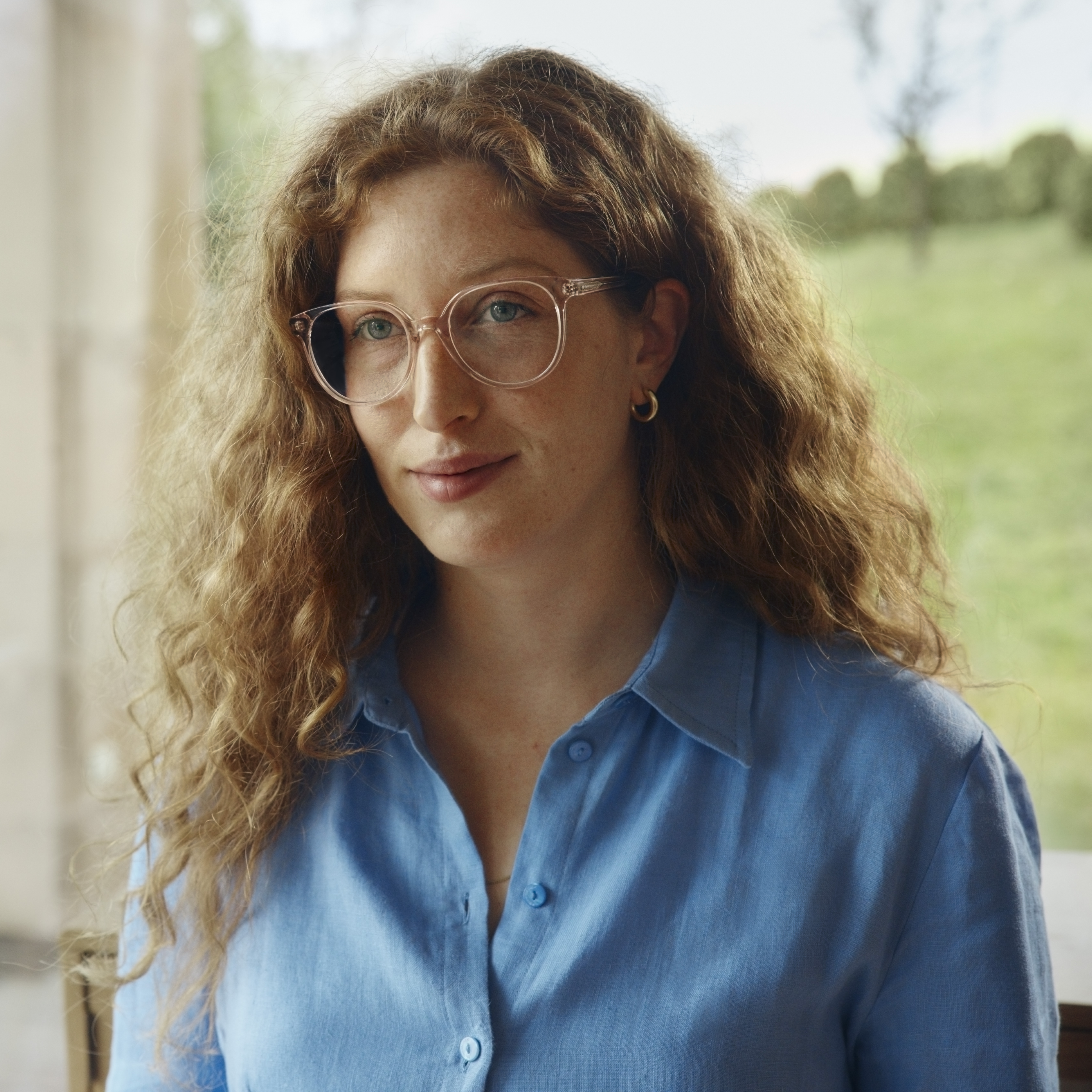 Eine Person mit lockigem Haar und Brille trägt ein blaues Hemd und steht in einem Raum vor einem Fenster, im Hintergrund ist eine grüne Landschaft zu sehen.