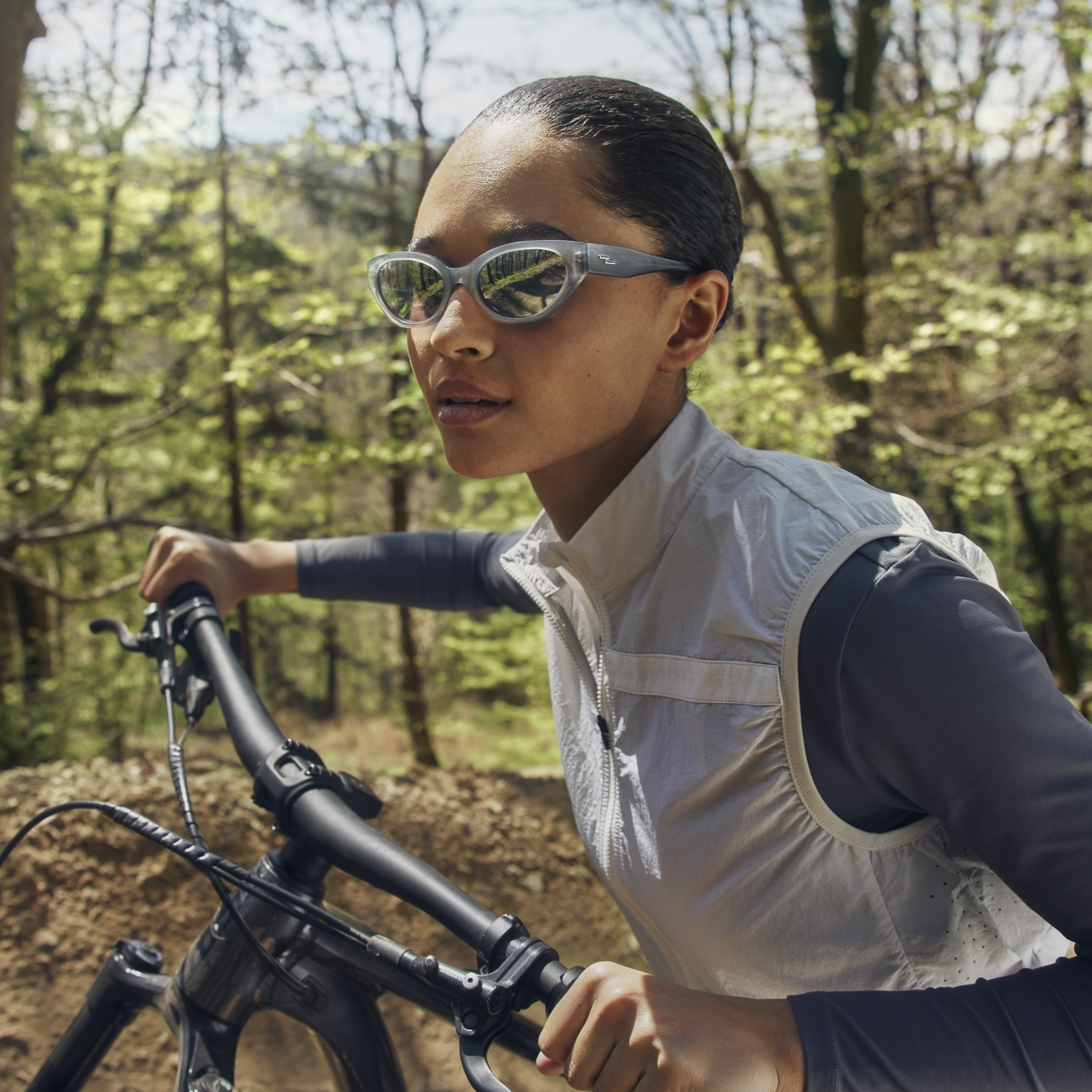 Eine Frau mit Sonnenbrille und einem weißen Top fährt mit dem Fahrrad durch eine sonnige, bewaldete Gegend, umgeben von grünen Bäumen.