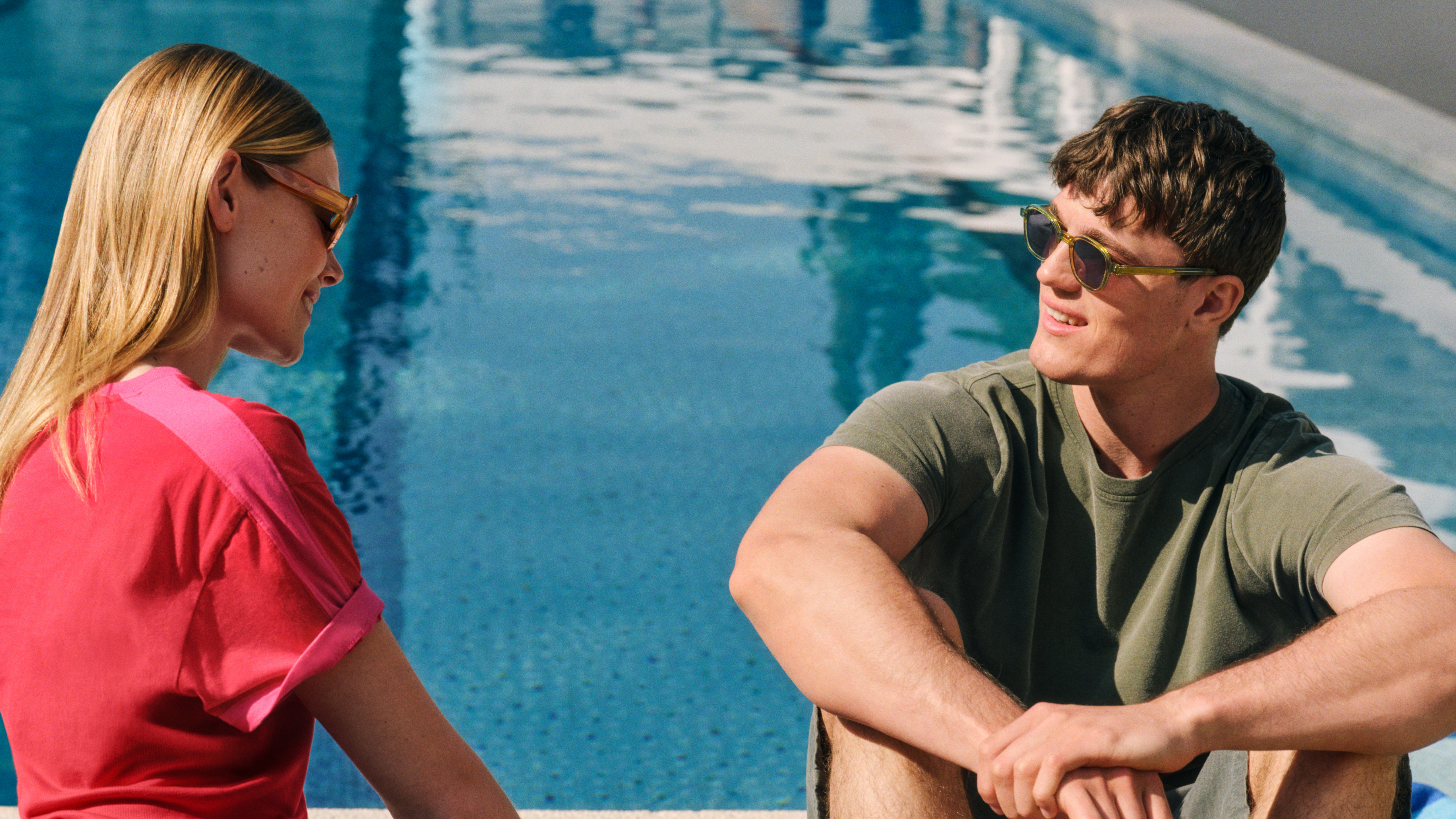 Zwei Menschen mit Sonnenbrillen sitzen am Pool, lächeln und unterhalten sich. Der eine trägt ein rosa Hemd, der andere ein grünes. Im Wasser spiegelt sich der Himmel.