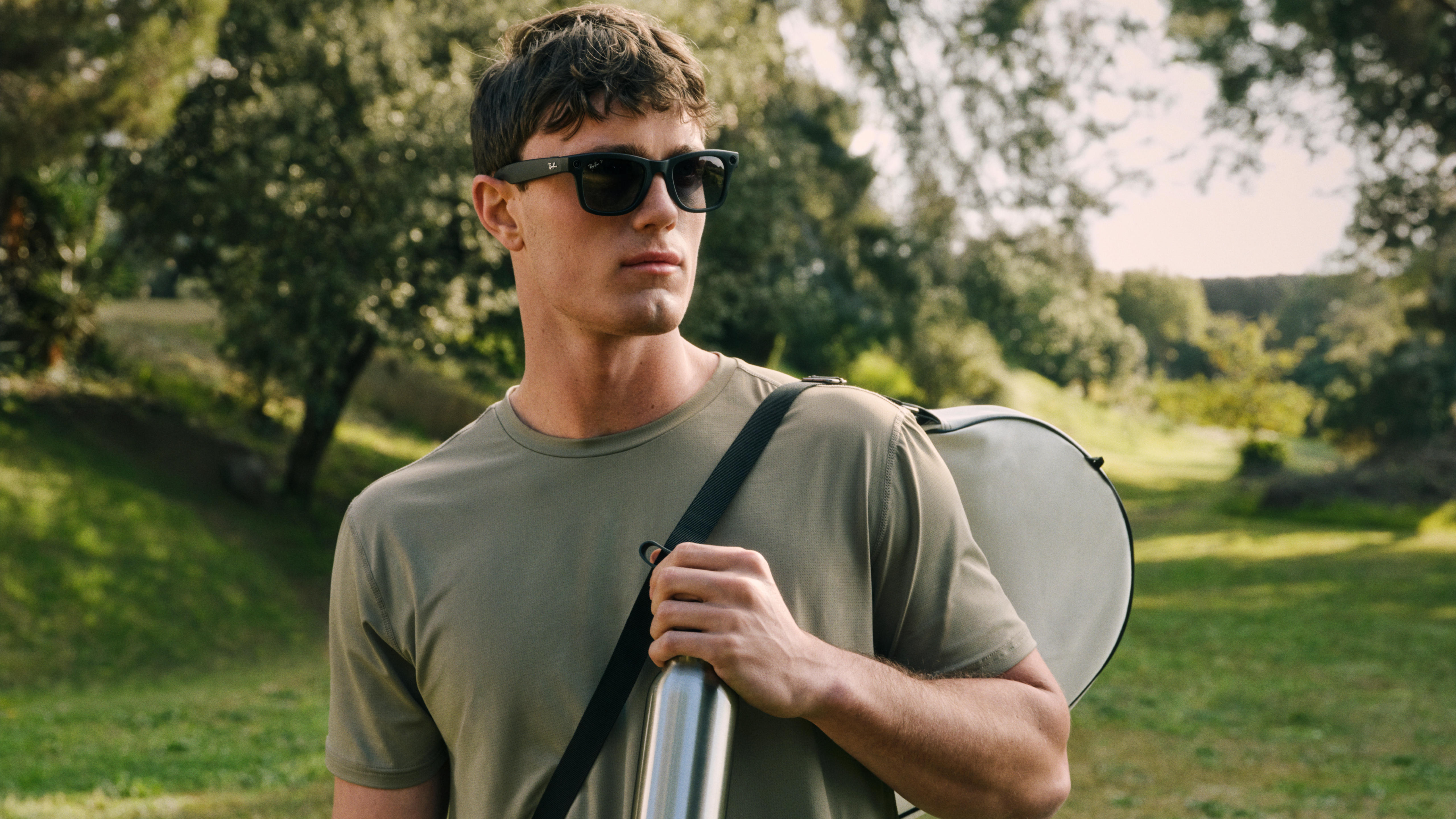 Ein Mann mit Sonnenbrille, der eine aufgerollte Yogamatte in der Hand hält, spaziert durch einen sonnigen Park, im Hintergrund sind Bäume und Gras zu sehen.