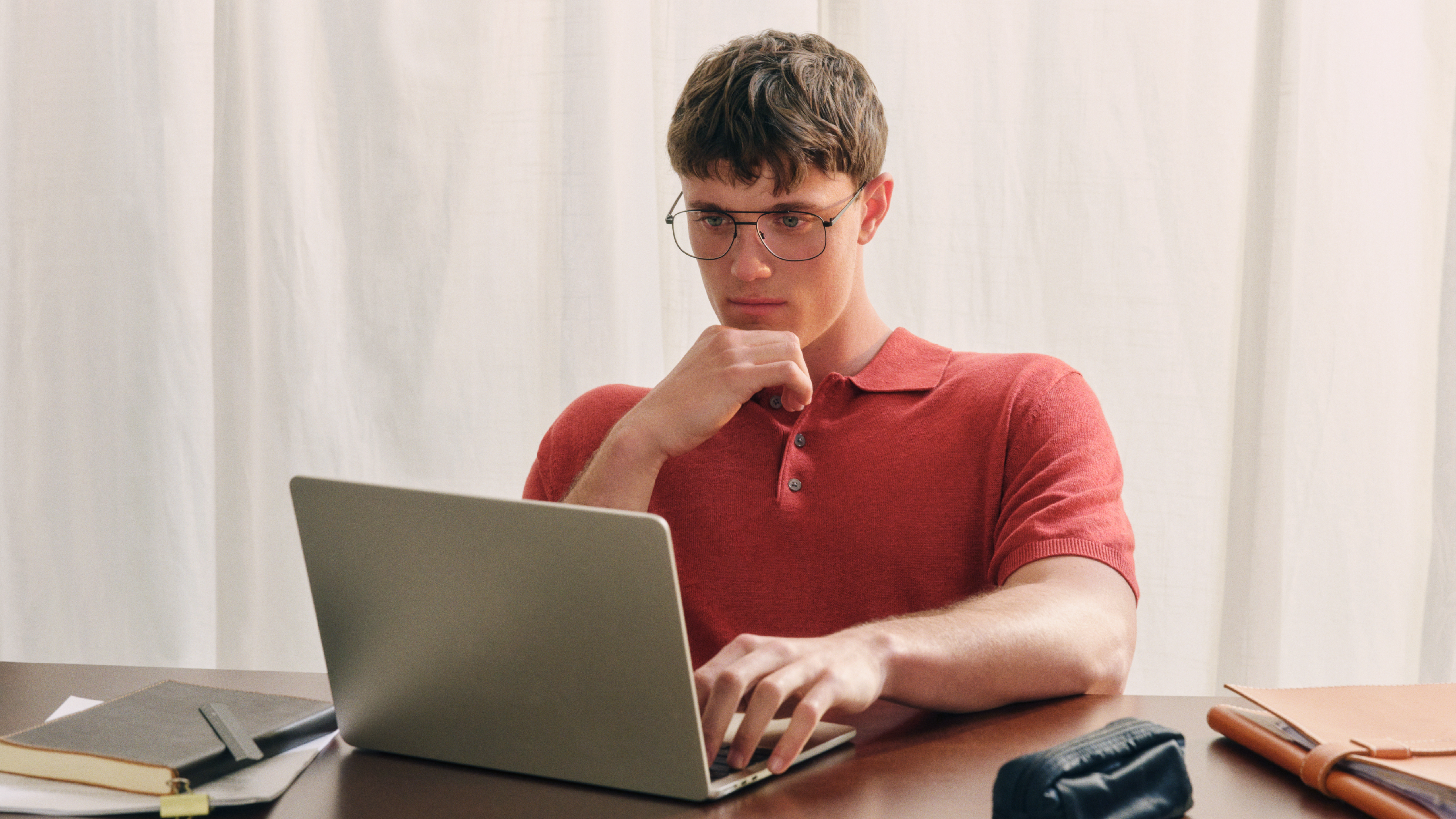 Eine Person mit Brille und rotem Polohemd arbeitet mit konzentriertem Gesichtsausdruck an einem Schreibtisch an einem Laptop, umgeben von Notizbüchern und einem Federmäppchen.
