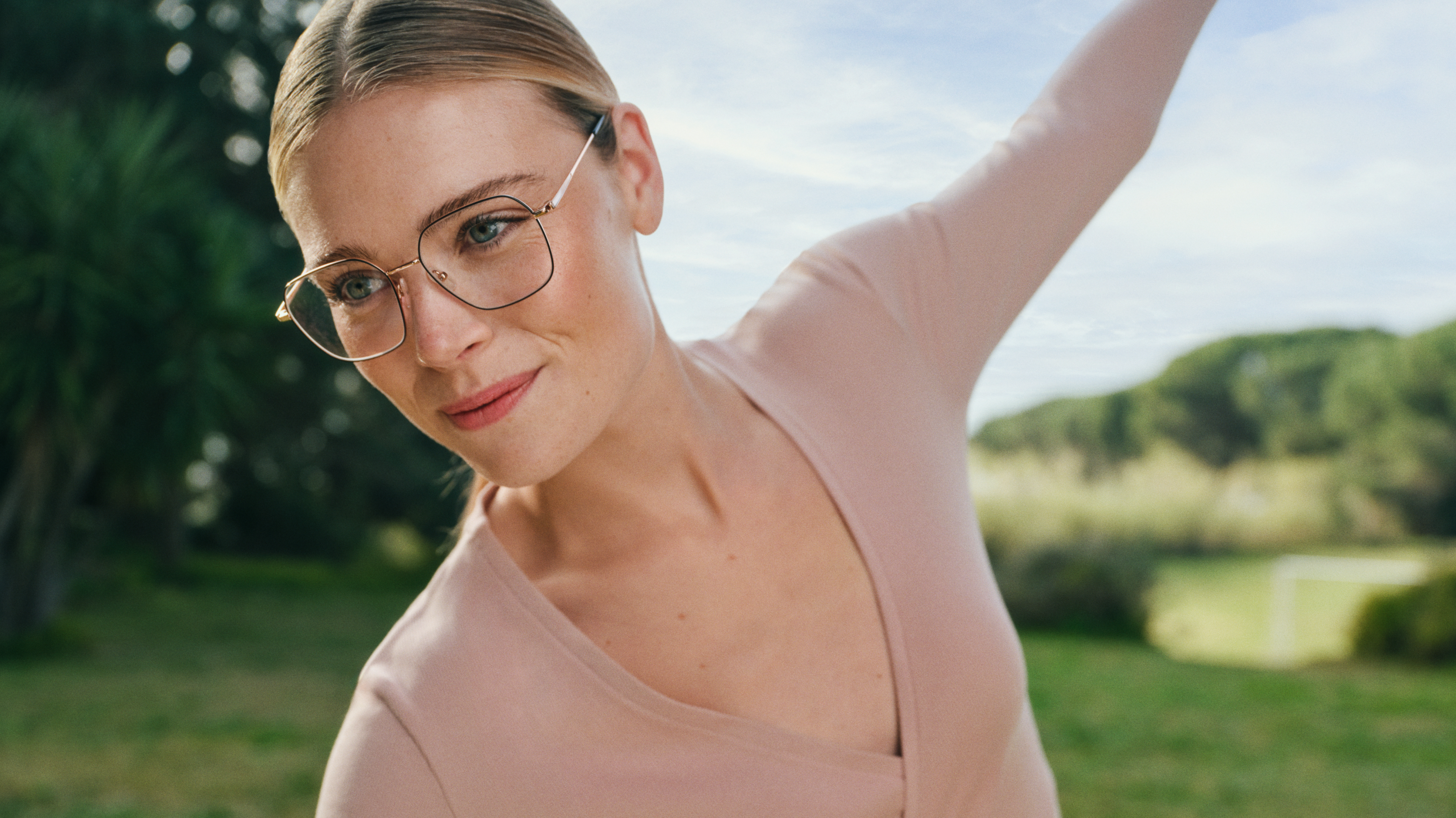 Eine Person mit Brille und einem hellrosa Oberteil macht draußen Dehnübungen, im Hintergrund sind Grünflächen und ein klarer Himmel zu sehen.