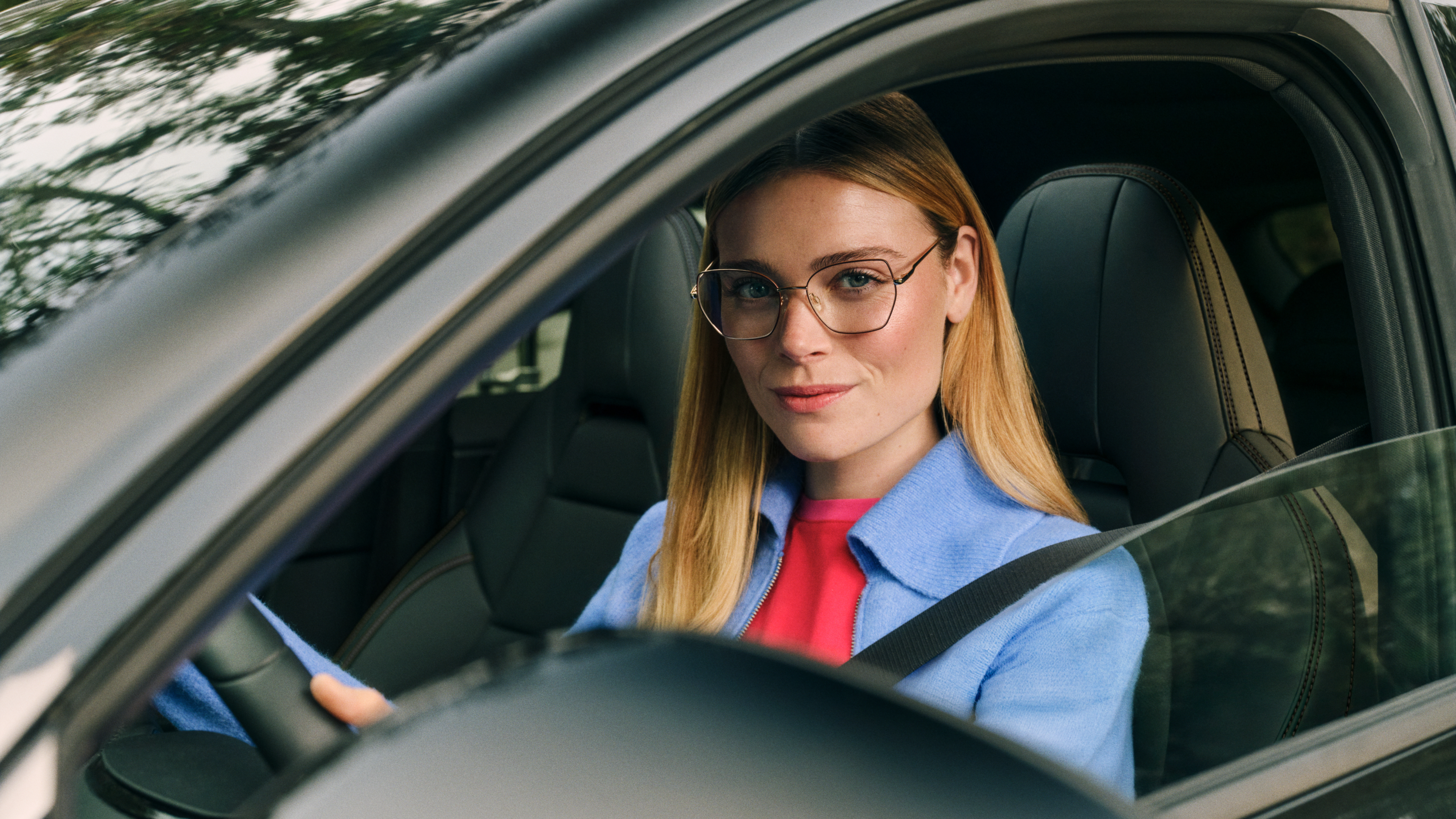Eine Frau mit Brille und blonden Haaren, die eine blaue Jacke trägt, sitzt auf dem Fahrersitz eines Autos, blickt direkt in die Kamera und ist von Bäumen umgeben.