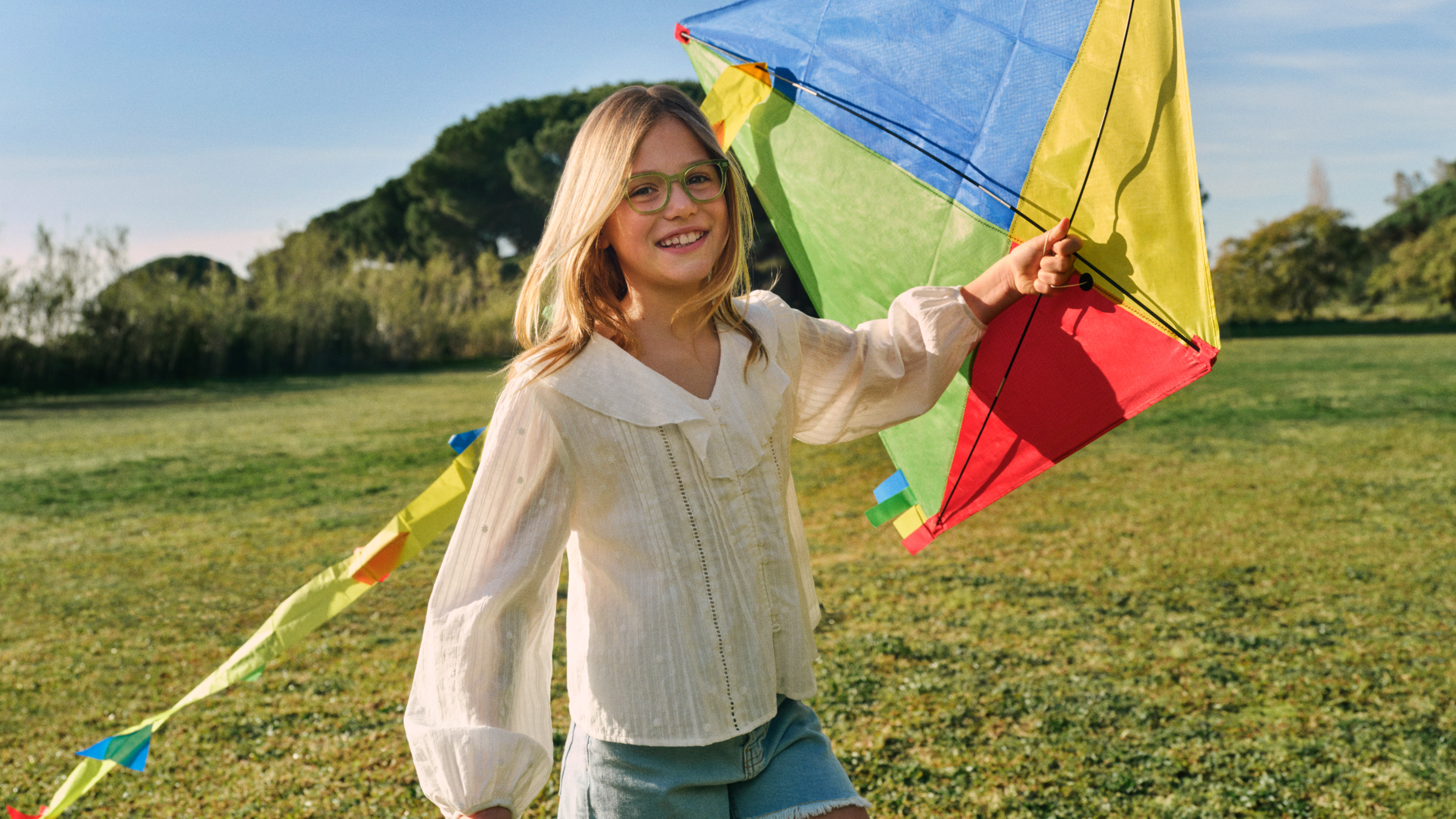 Ein lächelndes Mädchen mit Brille lässt auf einer sonnigen Wiese einen bunten Drachen steigen; sie trägt eine leichte Bluse und Shorts, im Hintergrund sind Bäume zu sehen.