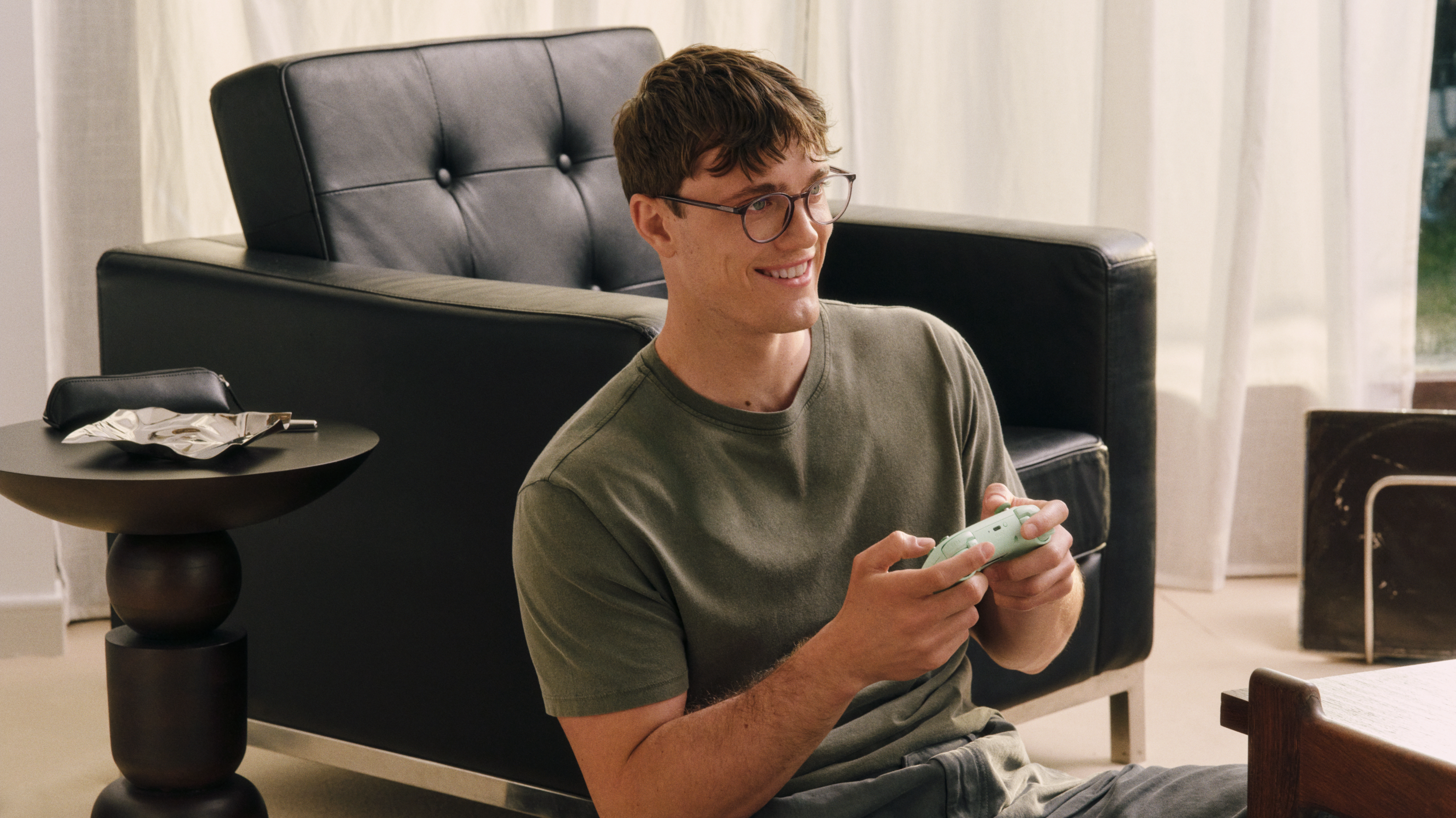 Un uomo con gli occhiali è seduto sul pavimento, sorride e tiene in mano un controller. Sullo sfondo si vedono una poltrona nera e un tavolino.