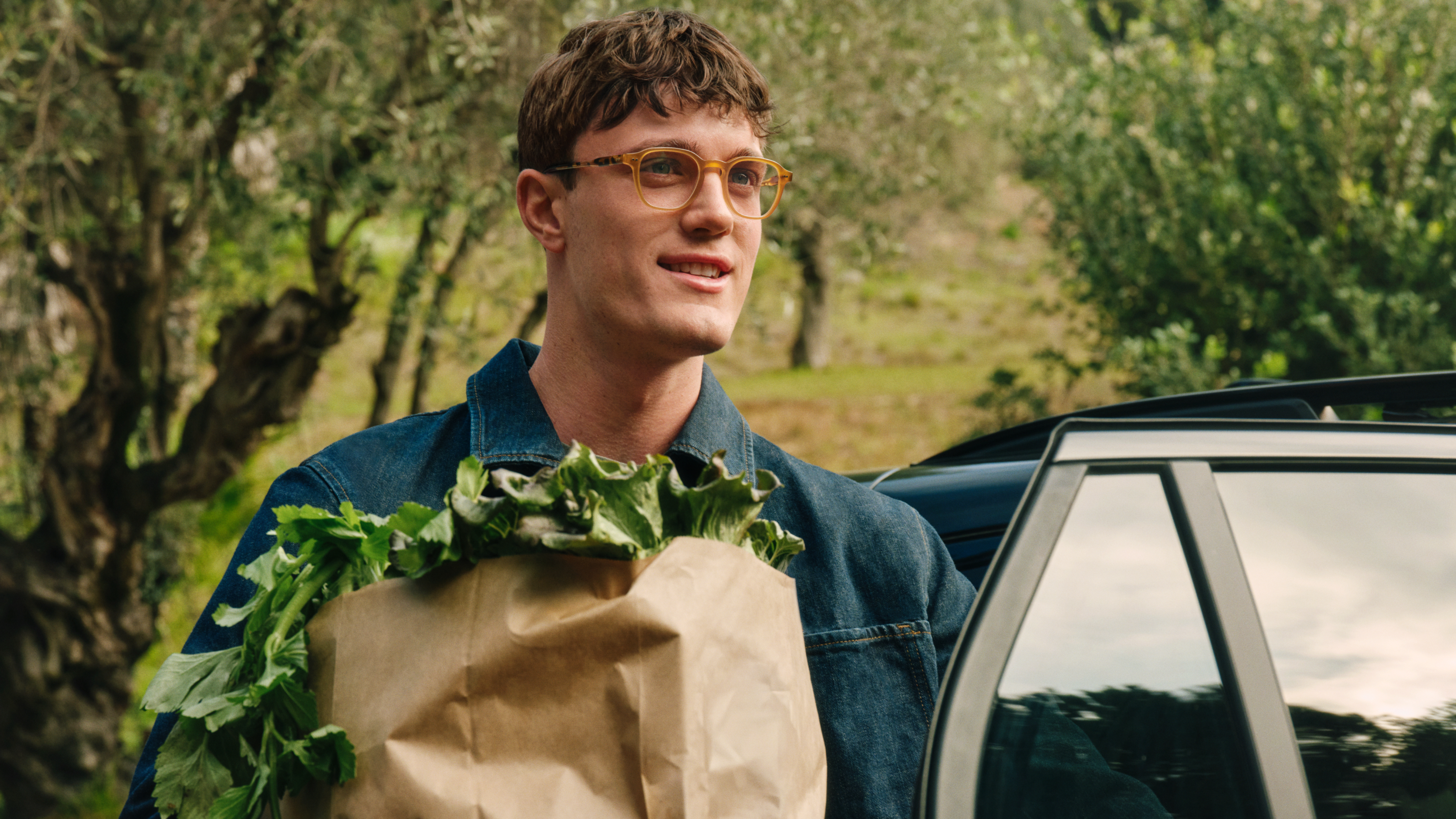 Ein Mann mit Brille lächelt, während er neben einem Auto eine mit Gemüse gefüllte Papiertüte in der Hand hält; im Hintergrund sind Bäume zu sehen.