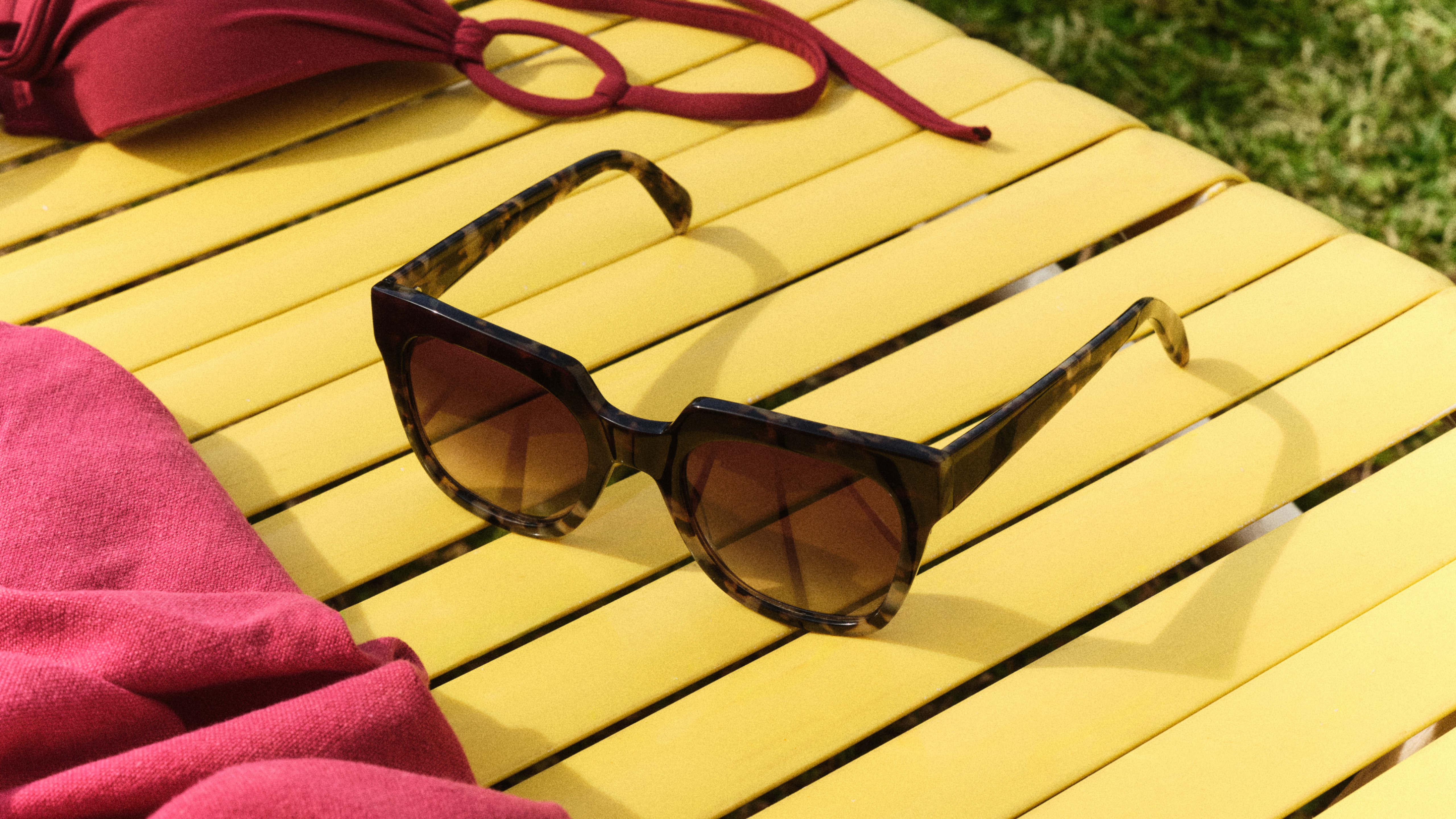 Eine Sonnenbrille in Schildpattfarbe auf einem gelben Tisch neben einem rosa Handtuch im Freien, wobei das Sonnenlicht Schatten wirft.