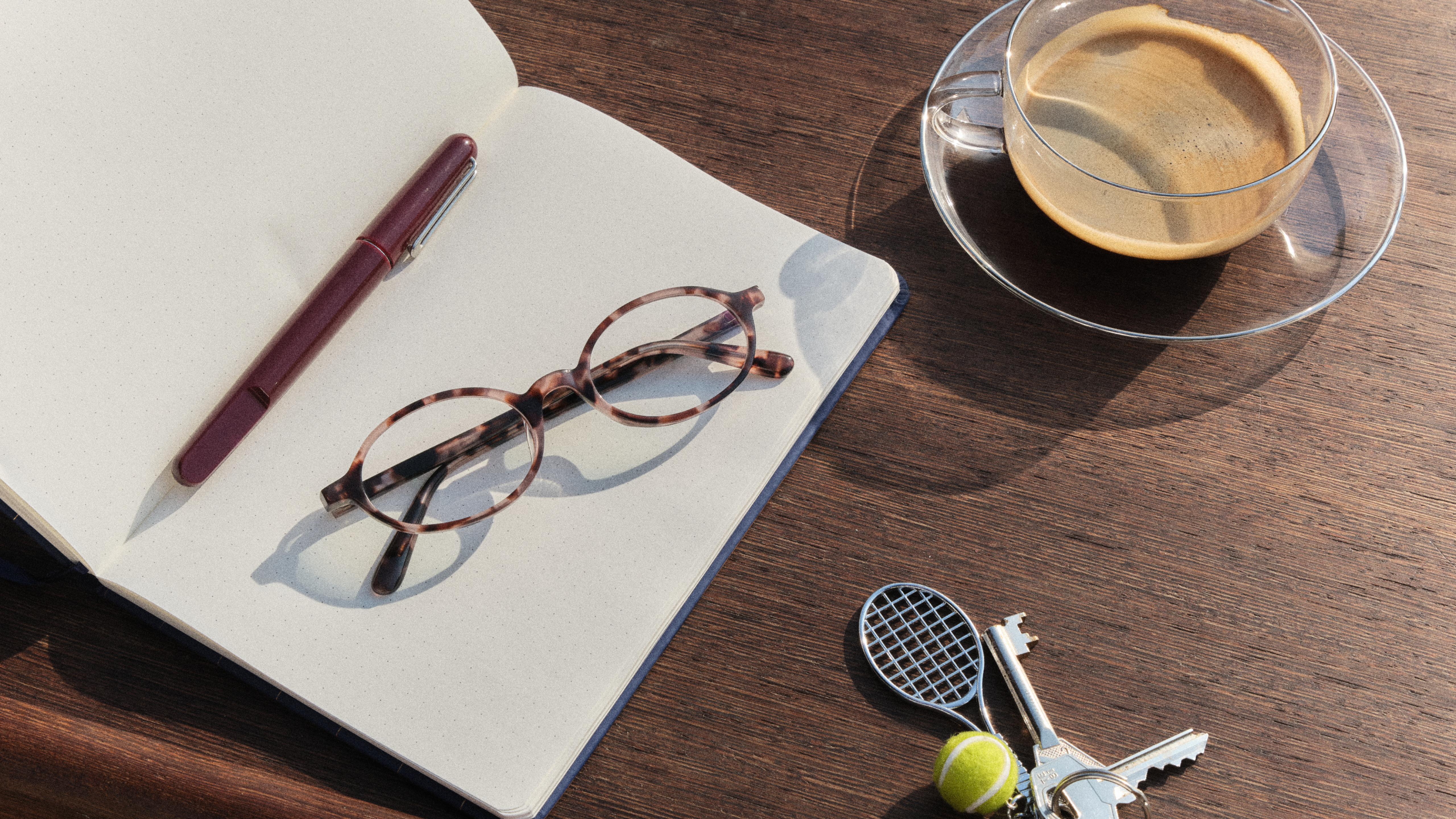 Otevřený notebook s brýlemi a perem, vedle něj cappuccino a přívěsek na klíče s miniaturní tenisovou raketou a míčkem na dřevěném stole.