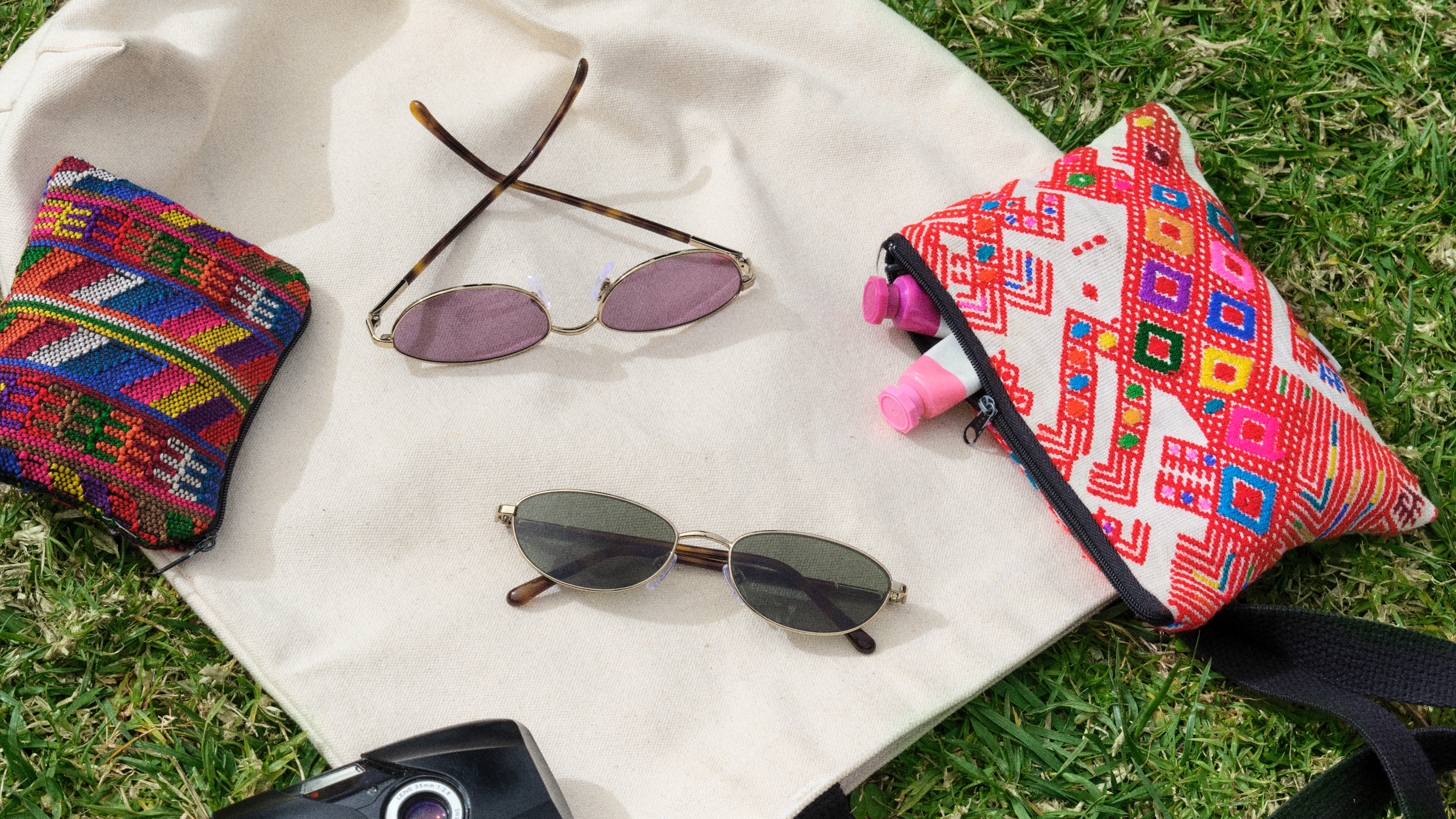 Außenaufnahme mit zwei Sonnenbrillen auf einem weißen Tuch, bunten Beuteln, Sonnencremeflaschen und einer Kamera auf dem Rasen.