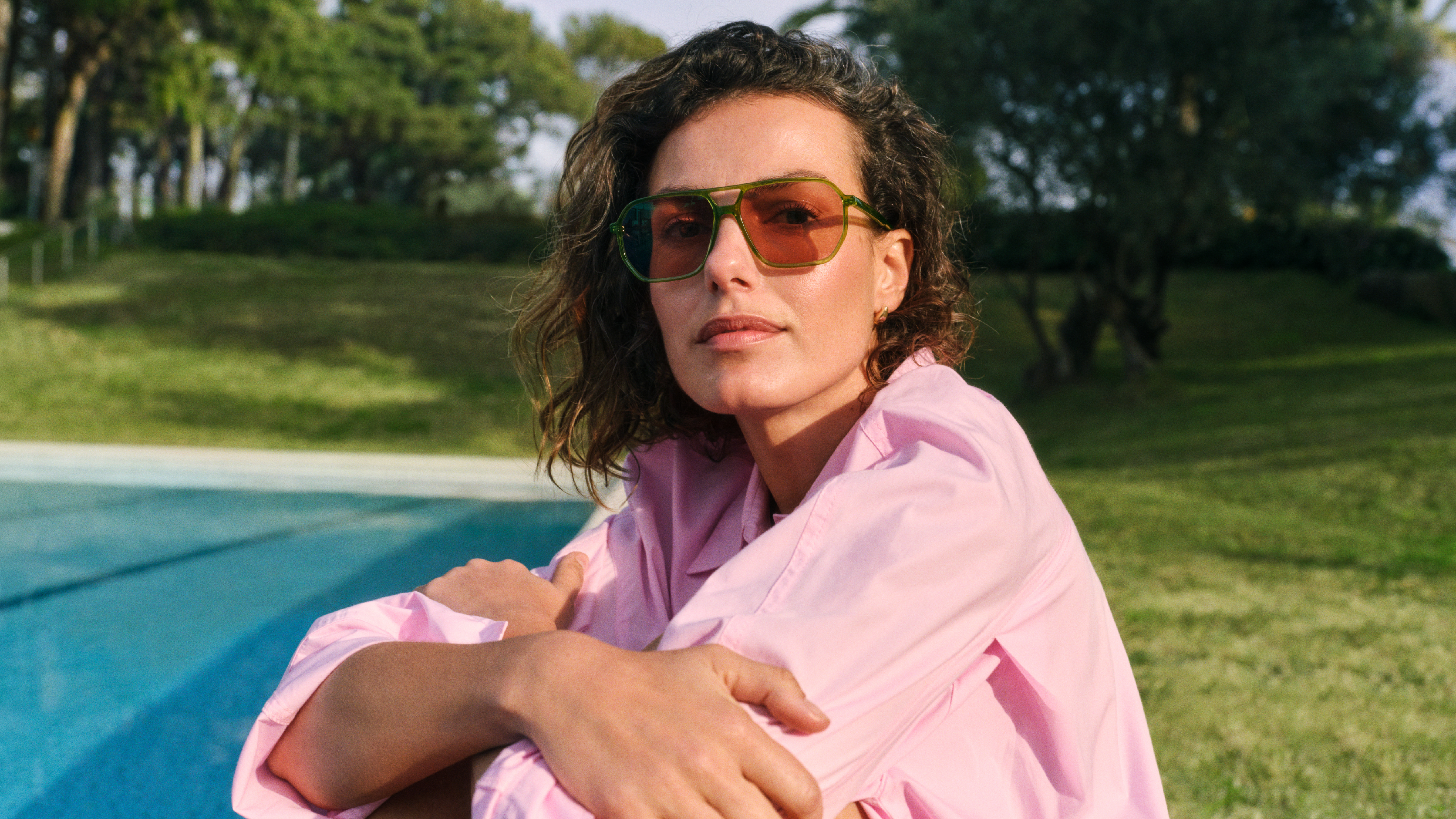 Eine Frau in einem rosa Shirt und mit Sonnenbrille entspannt sich am Pool, umgeben von einer grasbewachsenen Landschaft und Bäumen im Hintergrund.