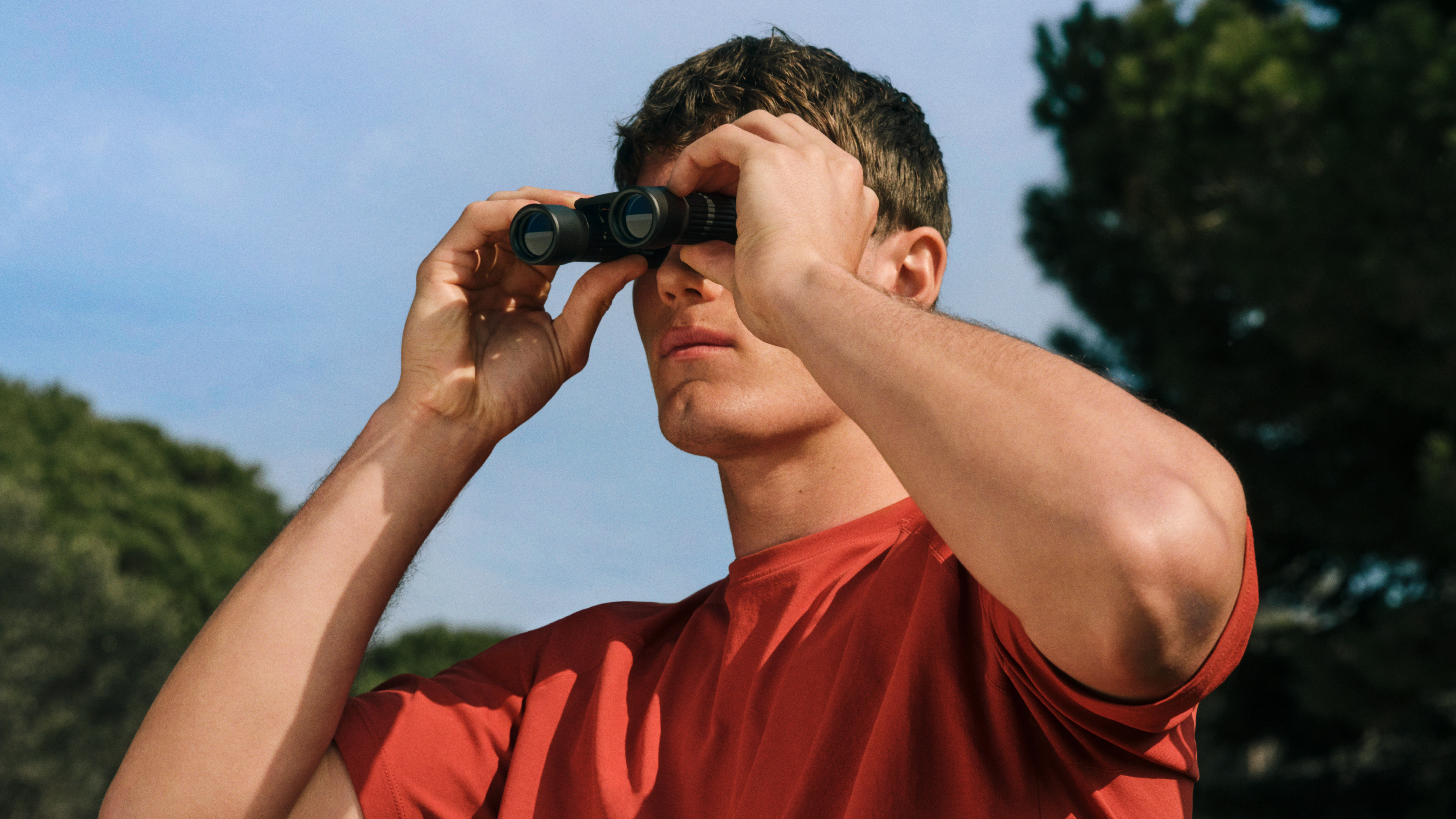 Una persona con una camicia rossa guarda attraverso un binocolo in una giornata soleggiata; sullo sfondo si vedono degli alberi e un cielo di un azzurro splendente.