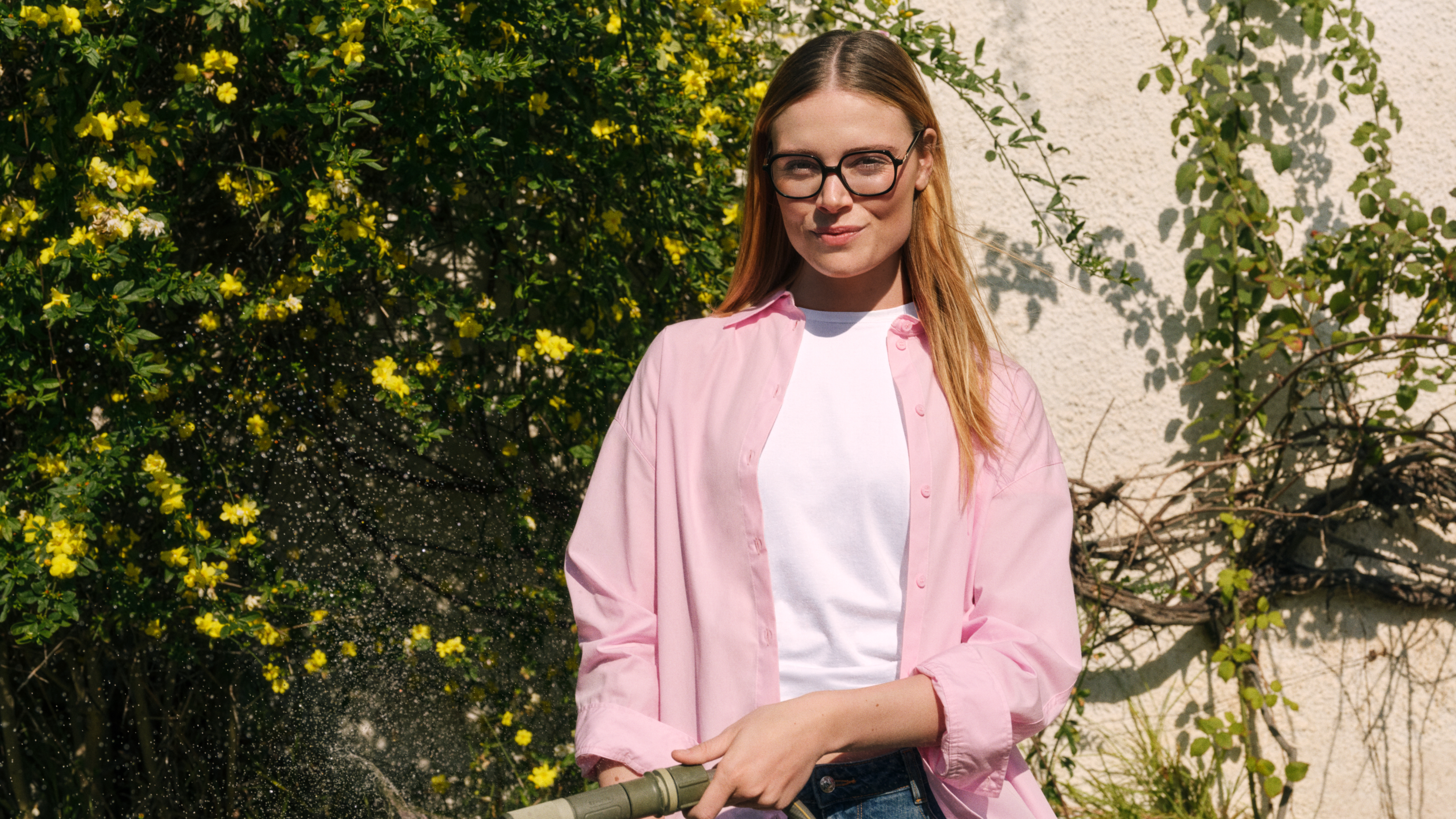 Eine Frau mit Brille und rosa Shirt, die in einem Garten mit einem Schlauch Pflanzen gießt und vor einem gelb blühenden Strauch und einer mit Weinreben bewachsenen Mauer steht.