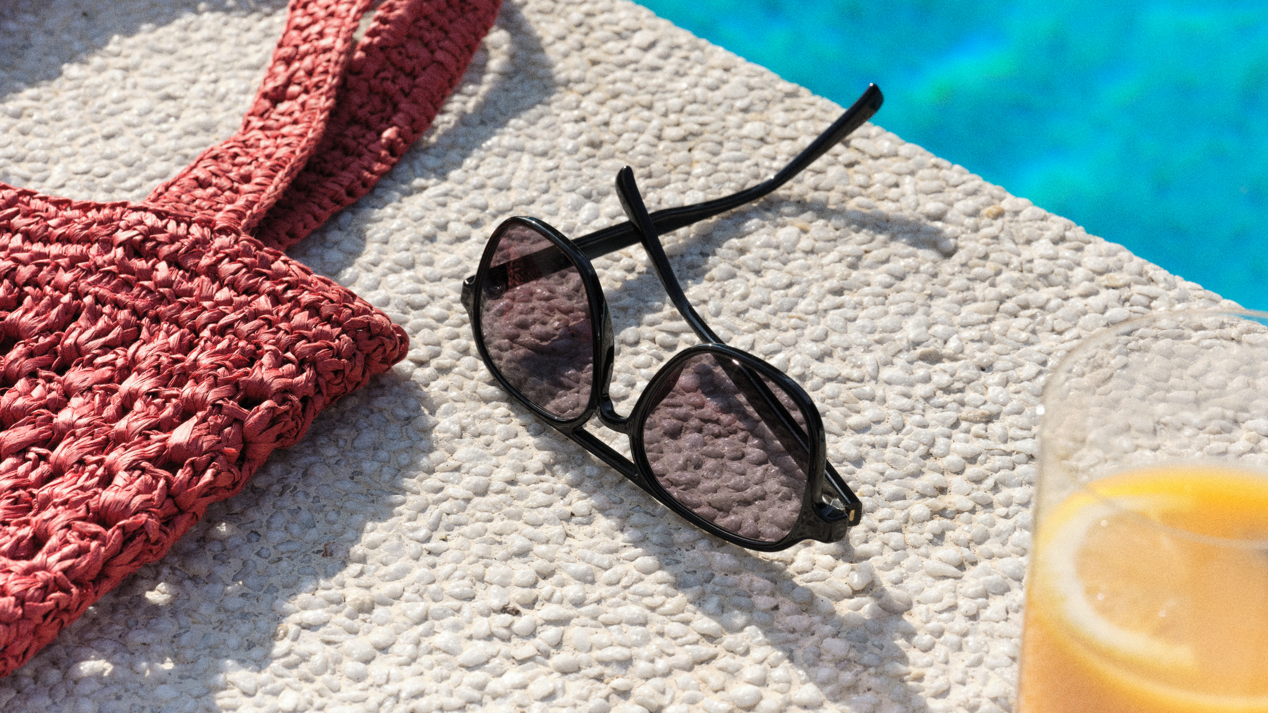 Une paire de lunettes de soleil posée sur une surface en pierre, à côté d'un sac en tissu rose et d'un verre de jus d'orange, près d'une piscine d'un bleu éclatant.