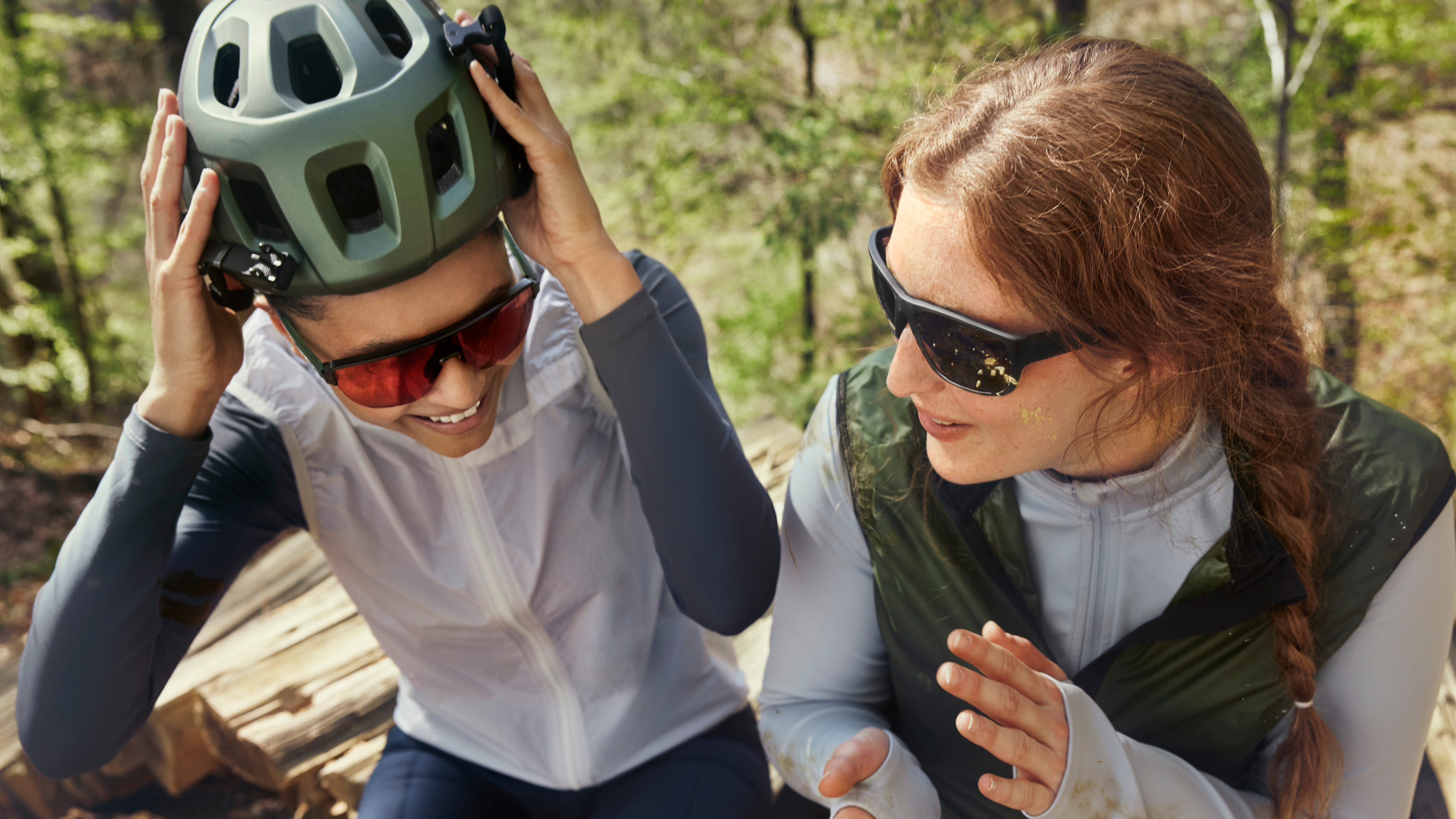 Due persone in abbigliamento da outdoor, una delle quali si sta sistemando il casco; entrambe sorridono e indossano occhiali da sole; sullo sfondo si vede un bosco.