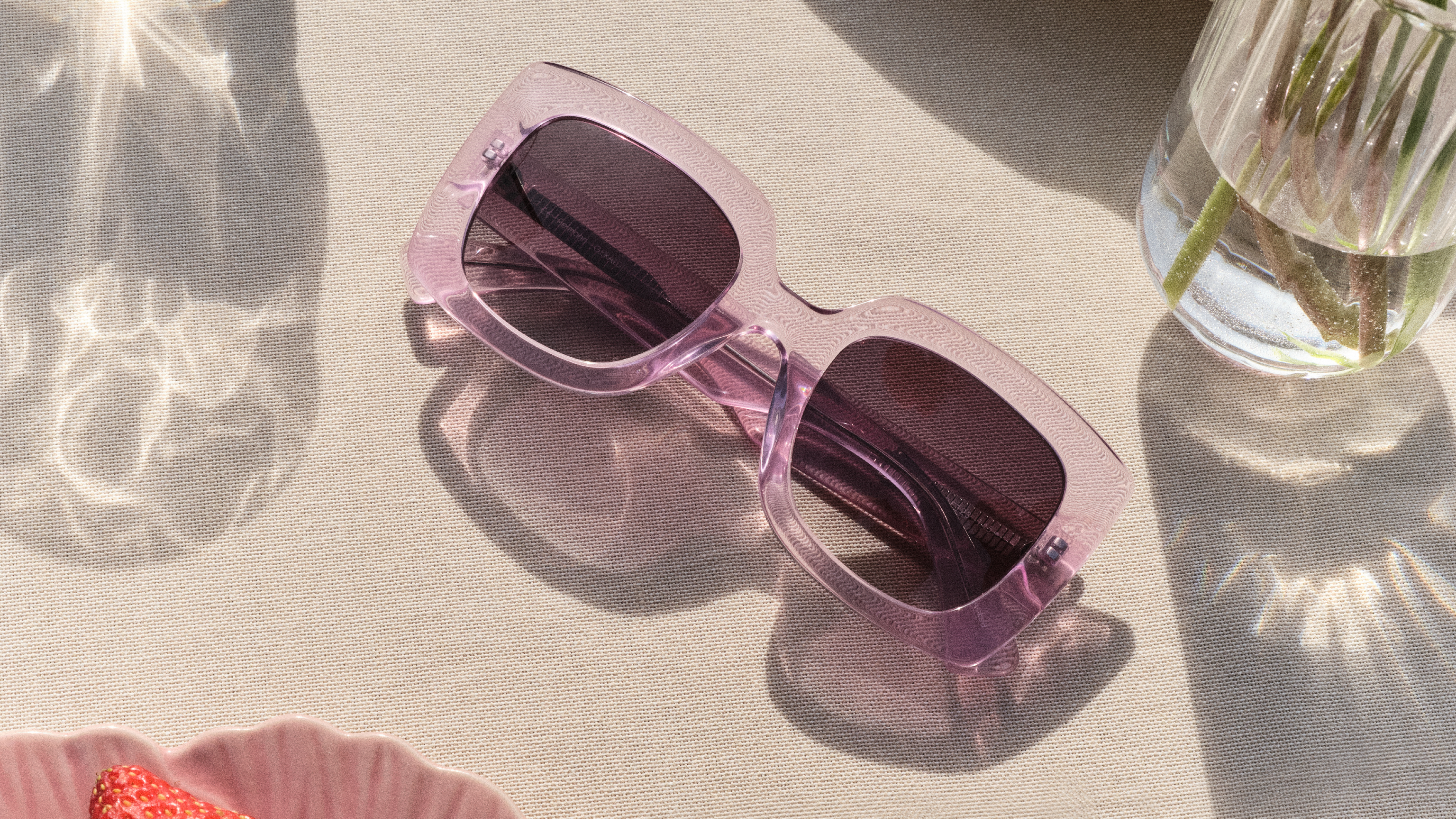 Eine rosa quadratische Sonnenbrille auf einer beigen Fläche, neben einer durchsichtigen Vase mit Wasser und einem rosa Teller, die sanfte Lichtreflexe erzeugt.