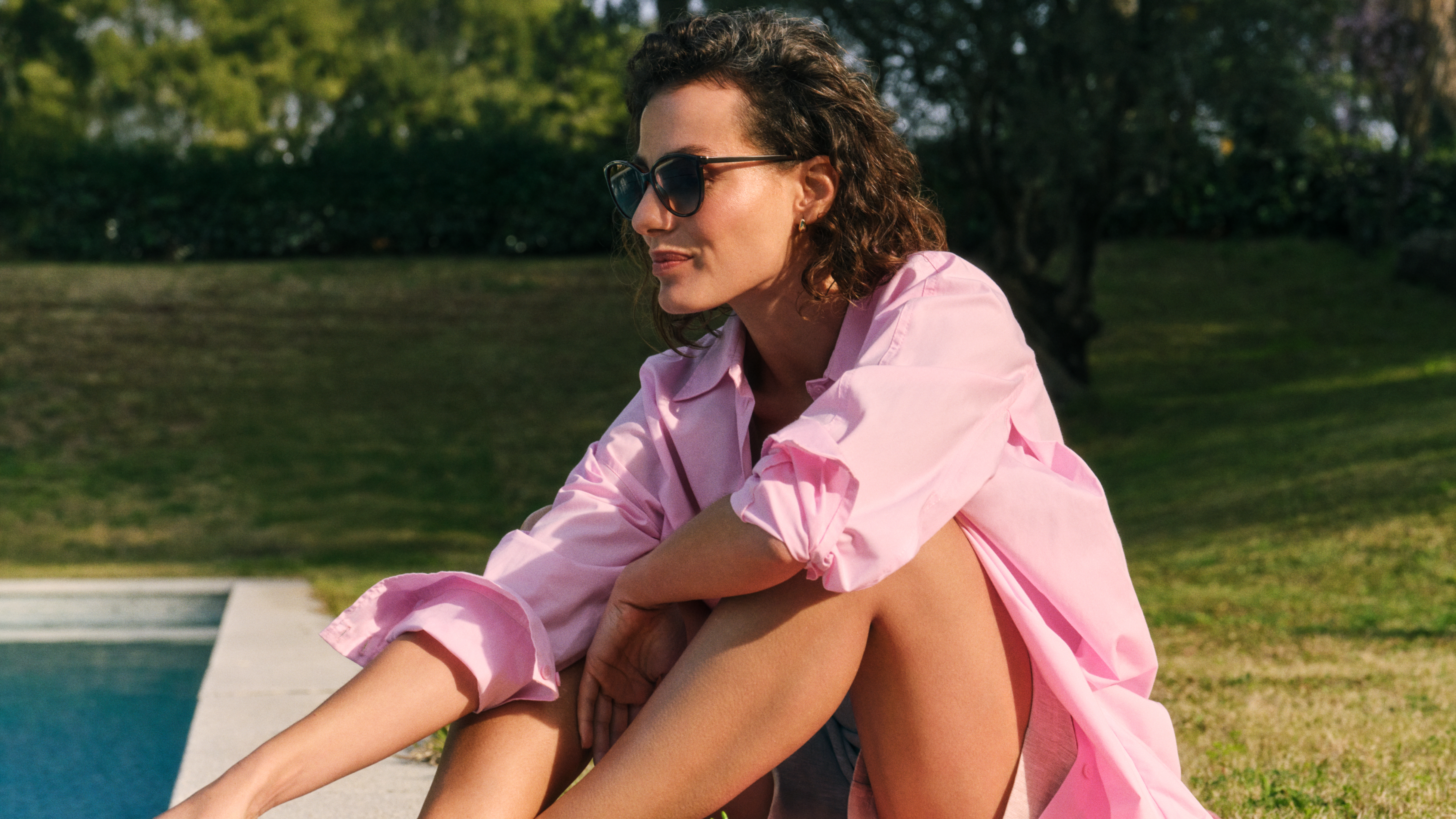 Eine Frau mit Sonnenbrille und rosa Shirt sitzt an einem sonnigen Tag draußen am Pool, im Hintergrund sind grünes Gras und Bäume zu sehen.
