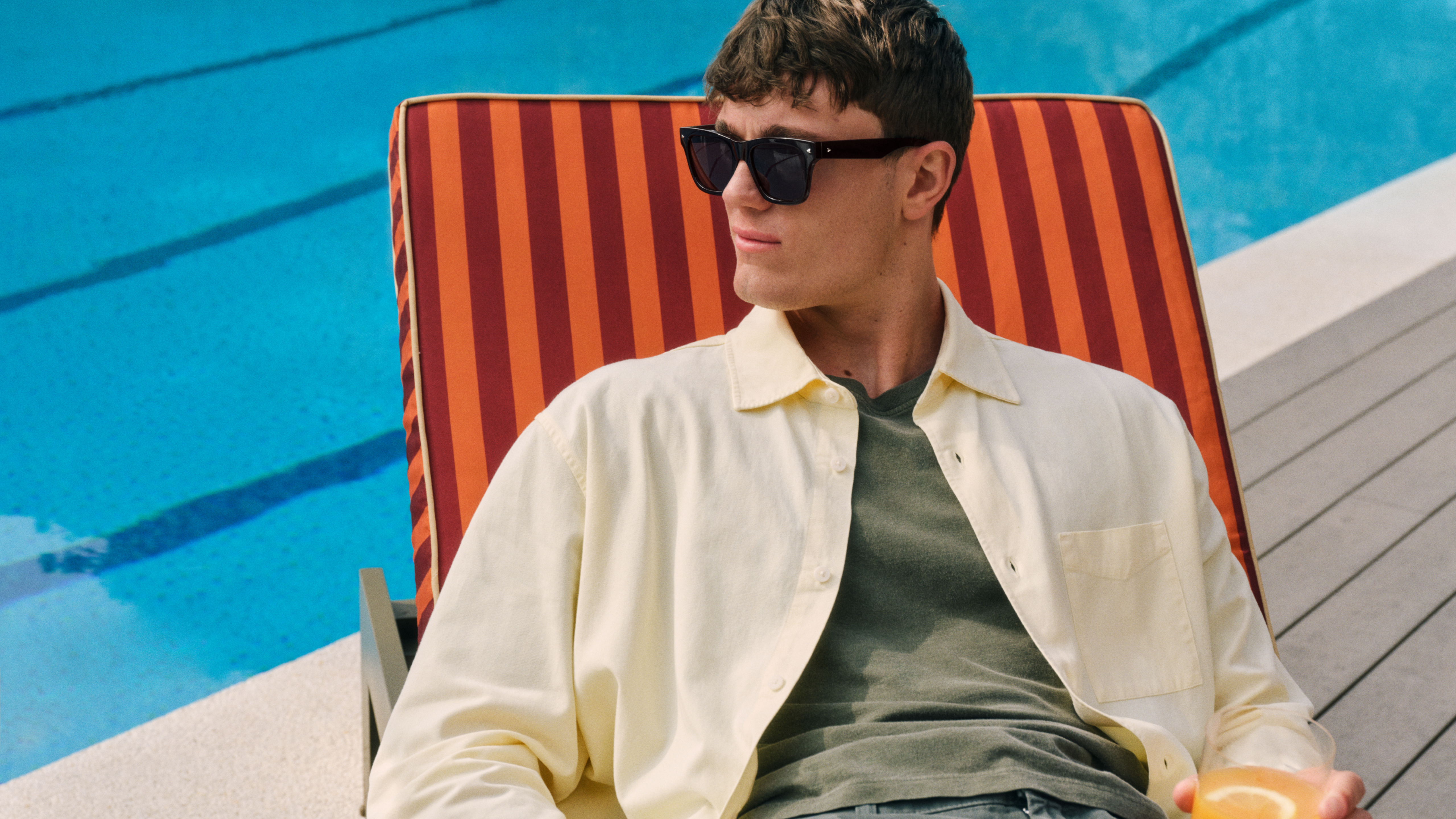 Un homme portant des lunettes de soleil, allongé dans une chaise longue rayée au bord de la piscine, vêtu d'une chemise claire et tenant une boisson jaune à la main.