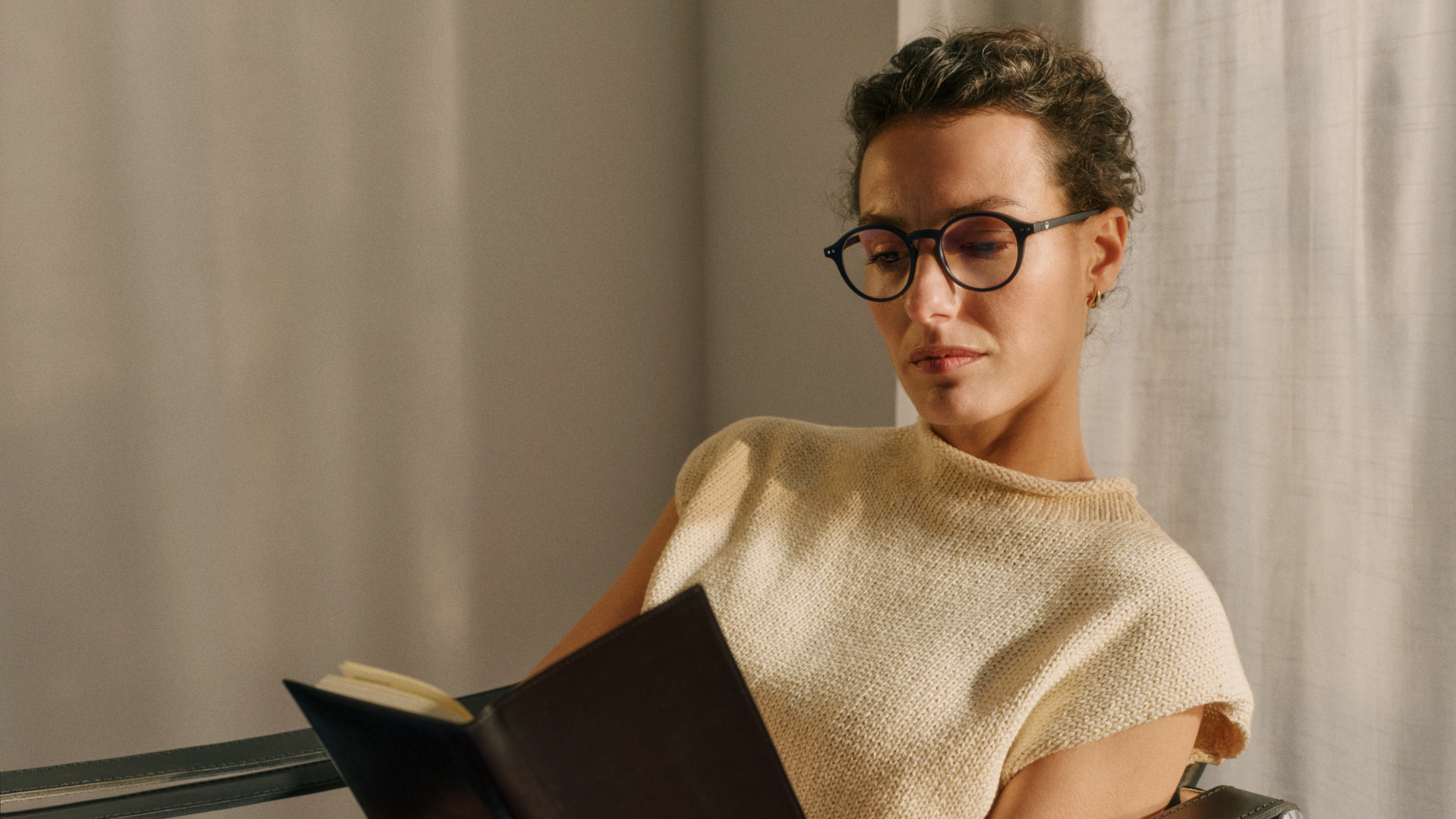 Eine Person mit Brille und kurzen Locken, die ein Buch liest, sitzt in einem Raum mit sanftem Licht und neutralen Farbtönen und trägt ein ärmelloses Oberteil.