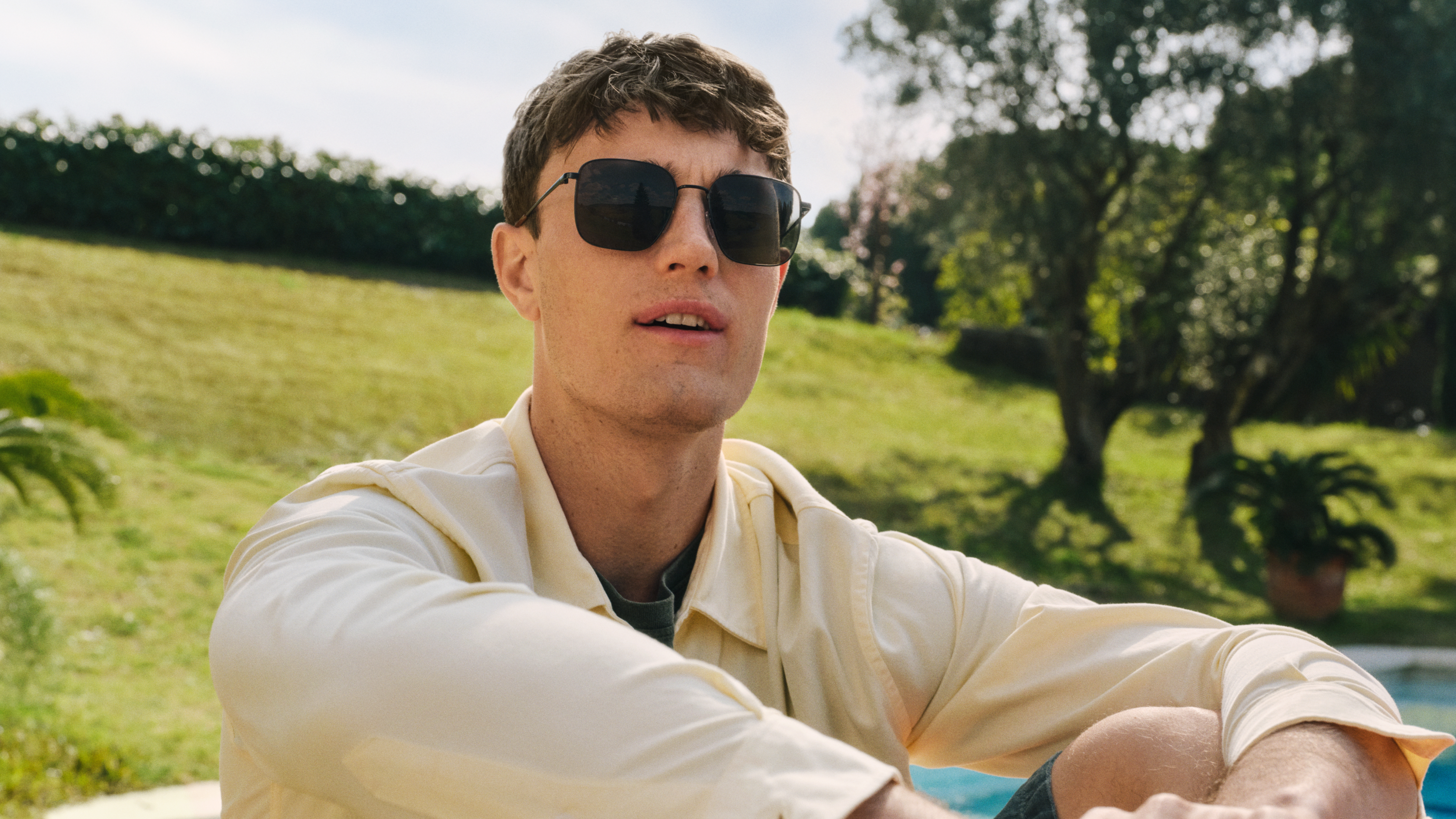 Un uomo è seduto all'aperto, indossa occhiali da sole e una camicia chiara. Si trova su un prato con alberi e una piscina sotto un cielo azzurro e limpido.