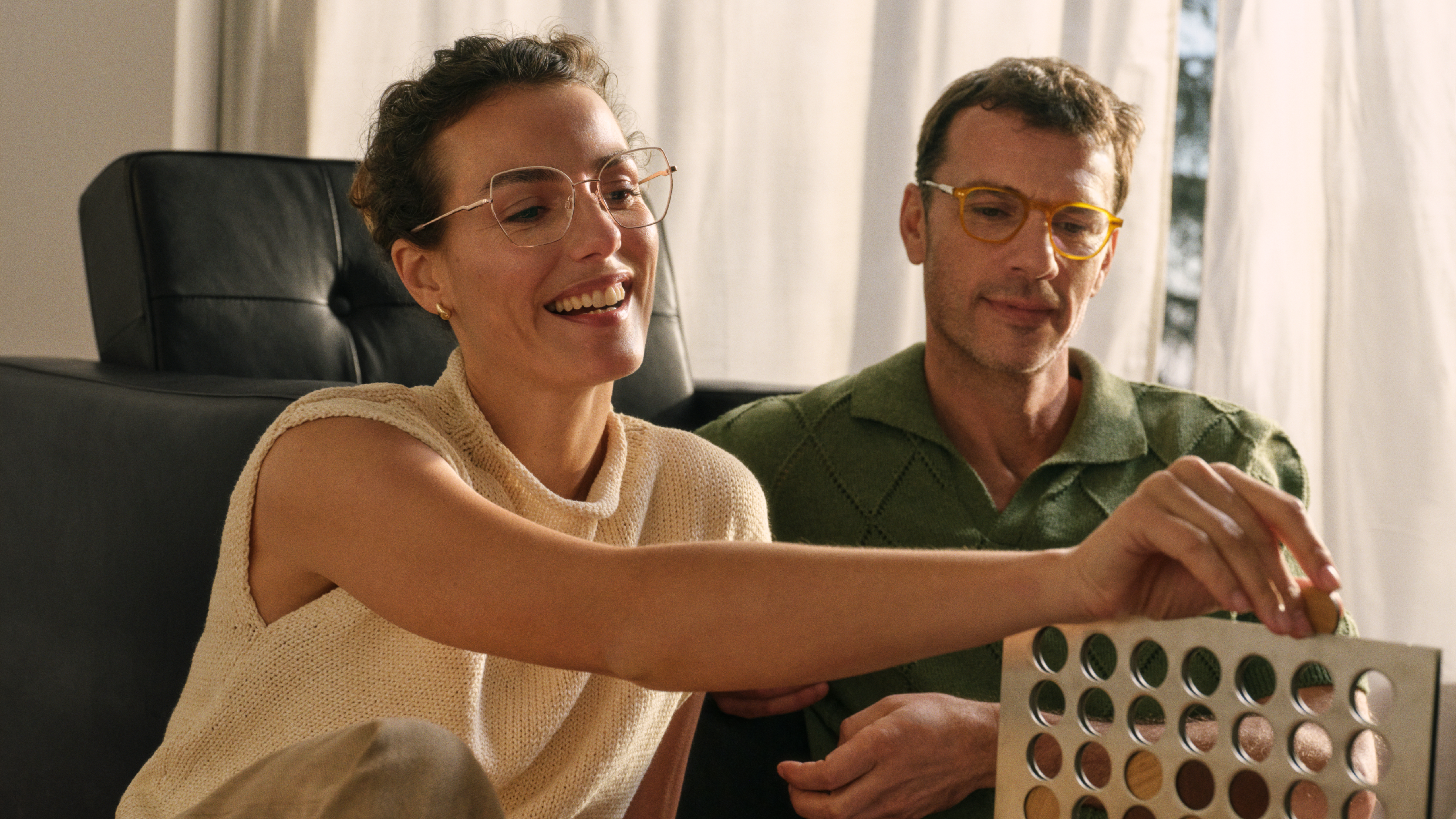 Eine Frau und ein Mann mit Brille spielen ein Spiel, lächeln und sitzen auf dem Boden in einem gemütlichen Raum mit sanfter Beleuchtung.