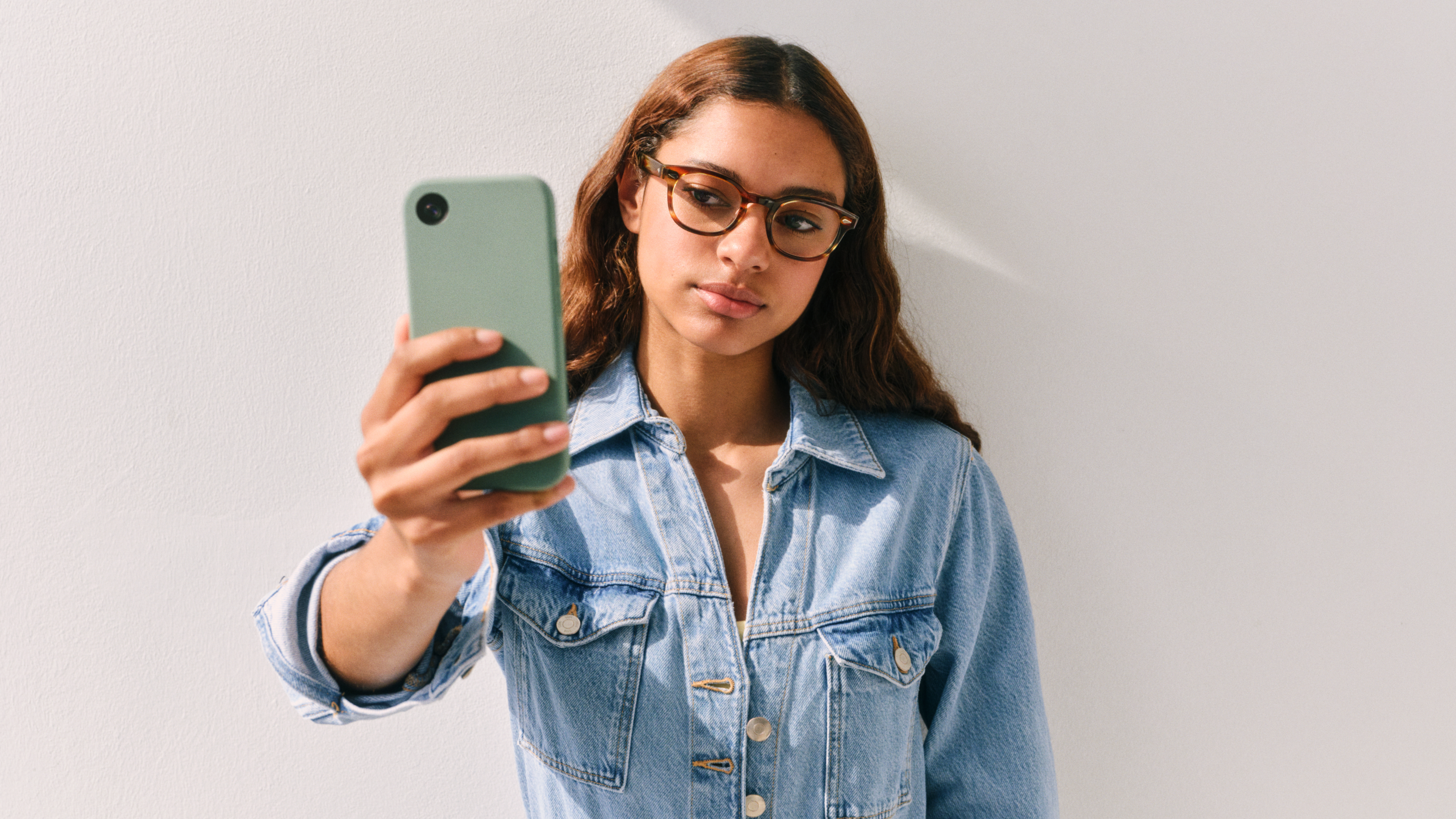 Eine Frau in Jeansjacke und Brille macht vor einer schlichten weißen Wand ein Selfie mit einem grünen Smartphone.