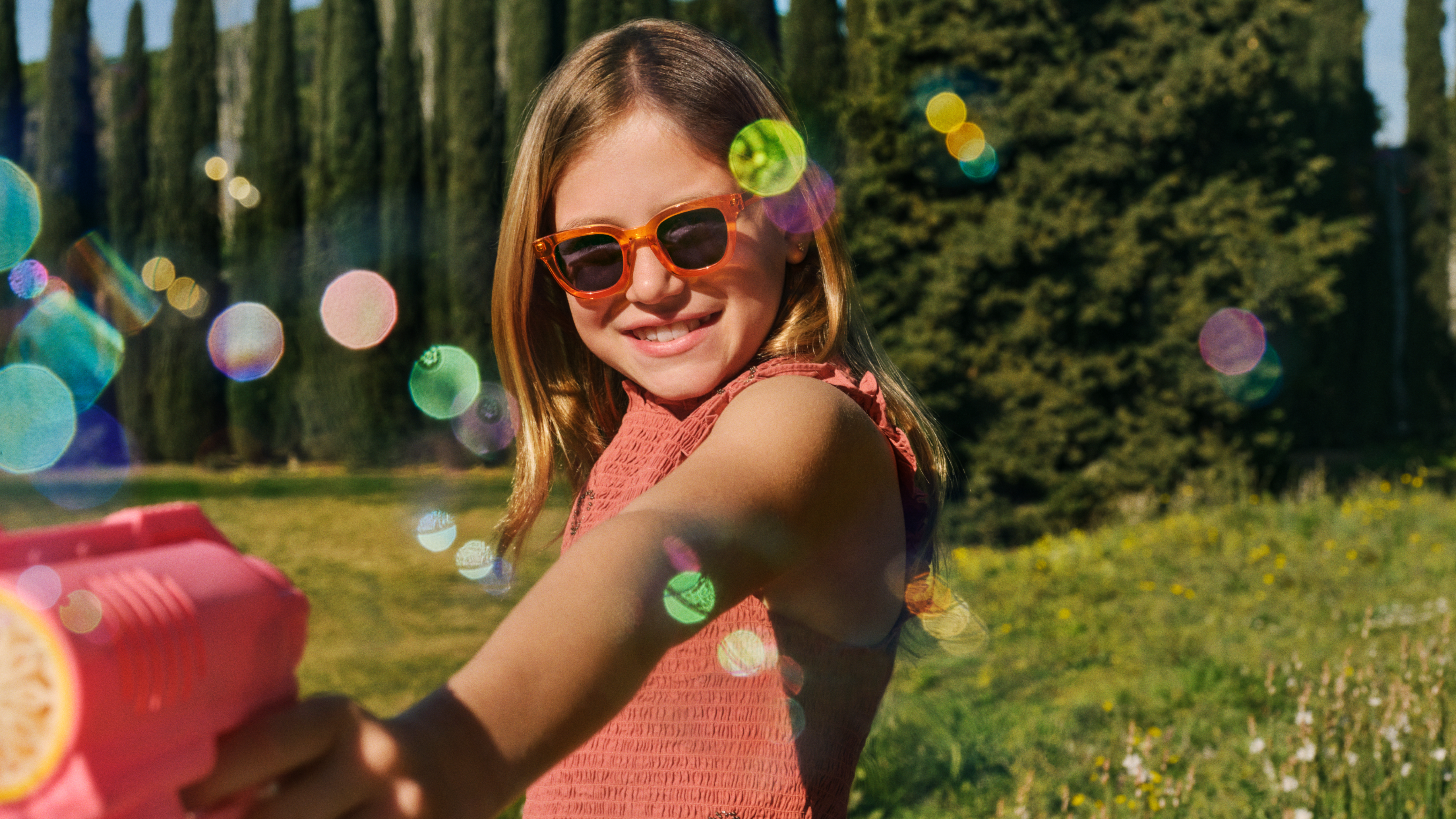 Una ragazzina con gli occhiali da sole sorride mentre tiene in mano una pistola ad acqua rosa in un parco soleggiato; sullo sfondo si vedono effetti di luce bokeh e cipressi.