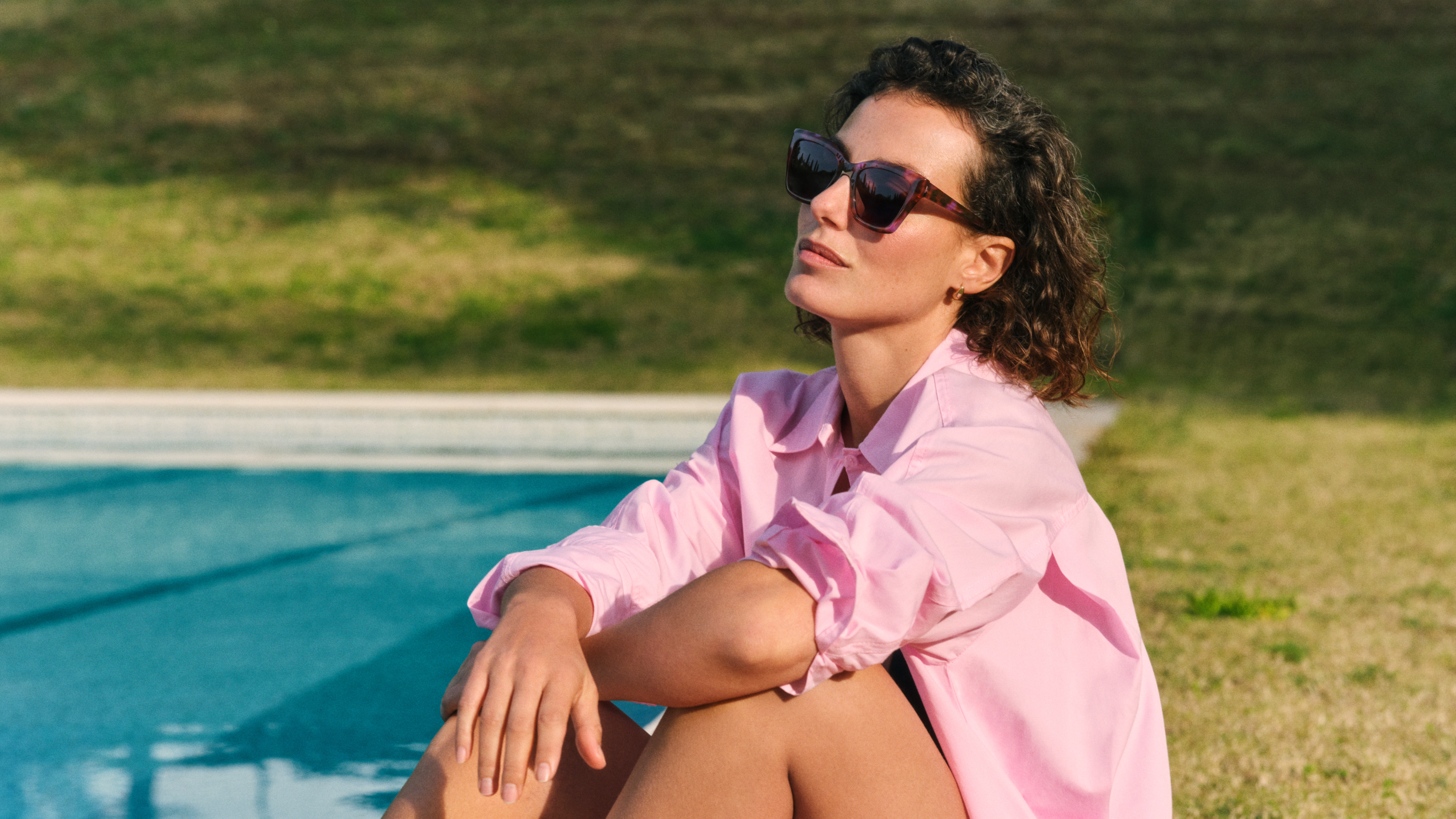 Kobieta w różowej koszulce i okularach przeciwsłonecznych siedzi przy basenie i zamyślona spogląda w dal. W tle widać trawę i odbicia słońca na wodzie.