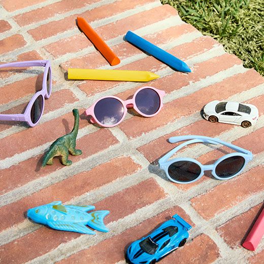 Drei Kindersonnenbrillen, Buntstifte, ein Spielzeugauto und ein kleiner Dinosaurier liegen auf einer Ziegeloberfläche verstreut.