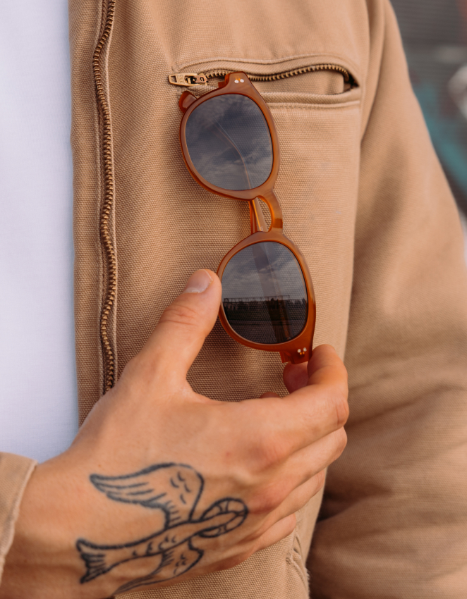Mano con un tatuaggio di un uccello che tiene degli occhiali da sole rotondi marroni attaccati alla tasca con cerniera di una giacca marrone chiaro.