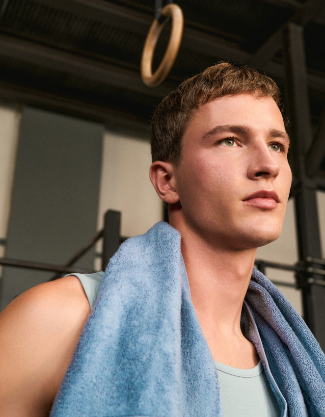 Eine Person mit kurzen Haaren und einem blauen Handtuch um den Hals steht in einer Turnhalle, im Hintergrund sind Turnringe zu sehen.